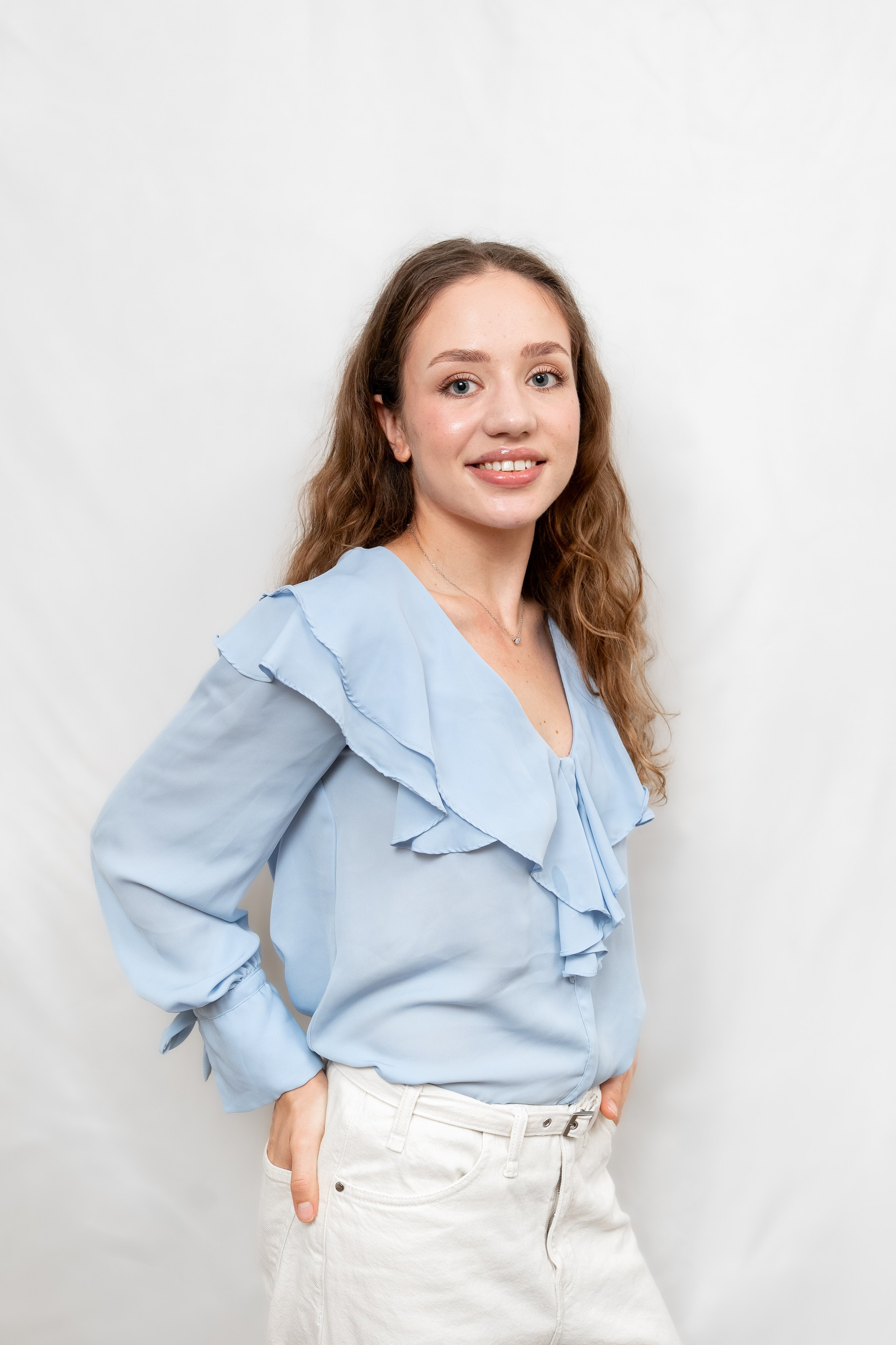 Confident young woman in a light blue blouse smiling with her hand in her pocket, standing against a white background.