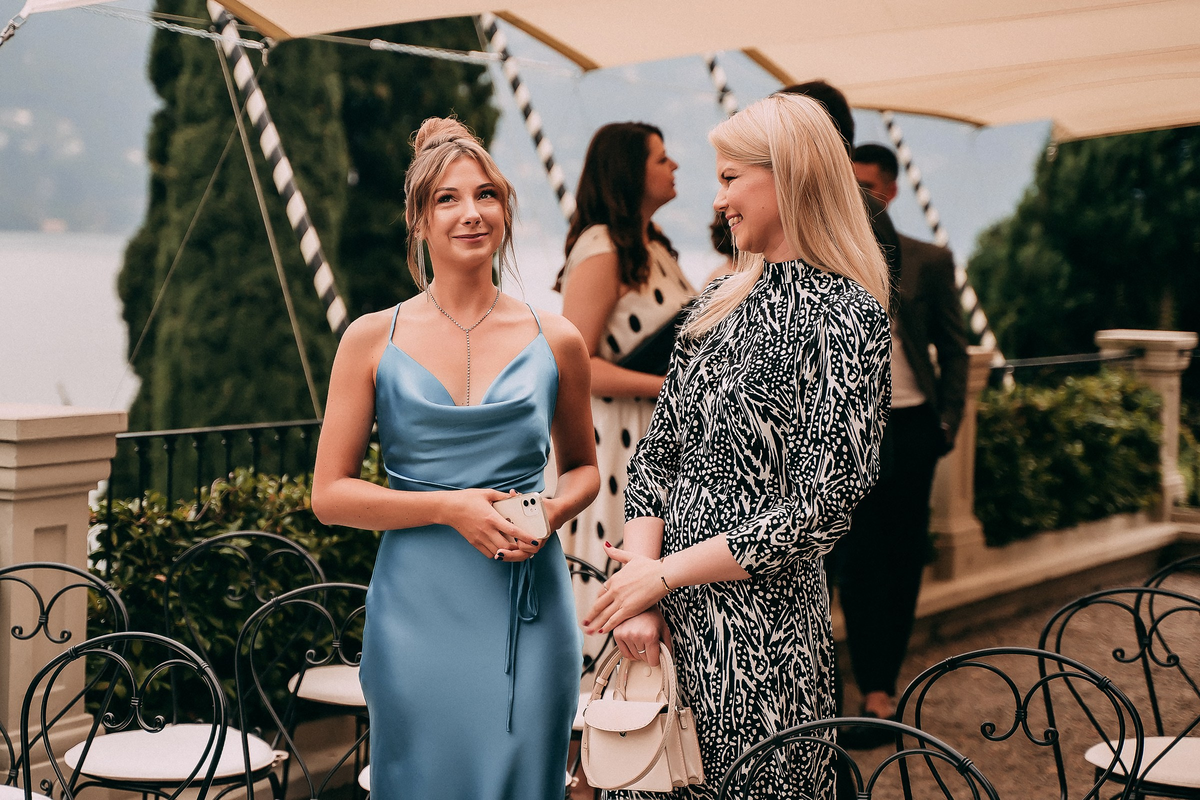 Two elegantly dressed women chatting at a sophisticated outdoor wedding reception by Lake Como, with classic black wrought-iron chairs and a stunning lakeside backdrop.