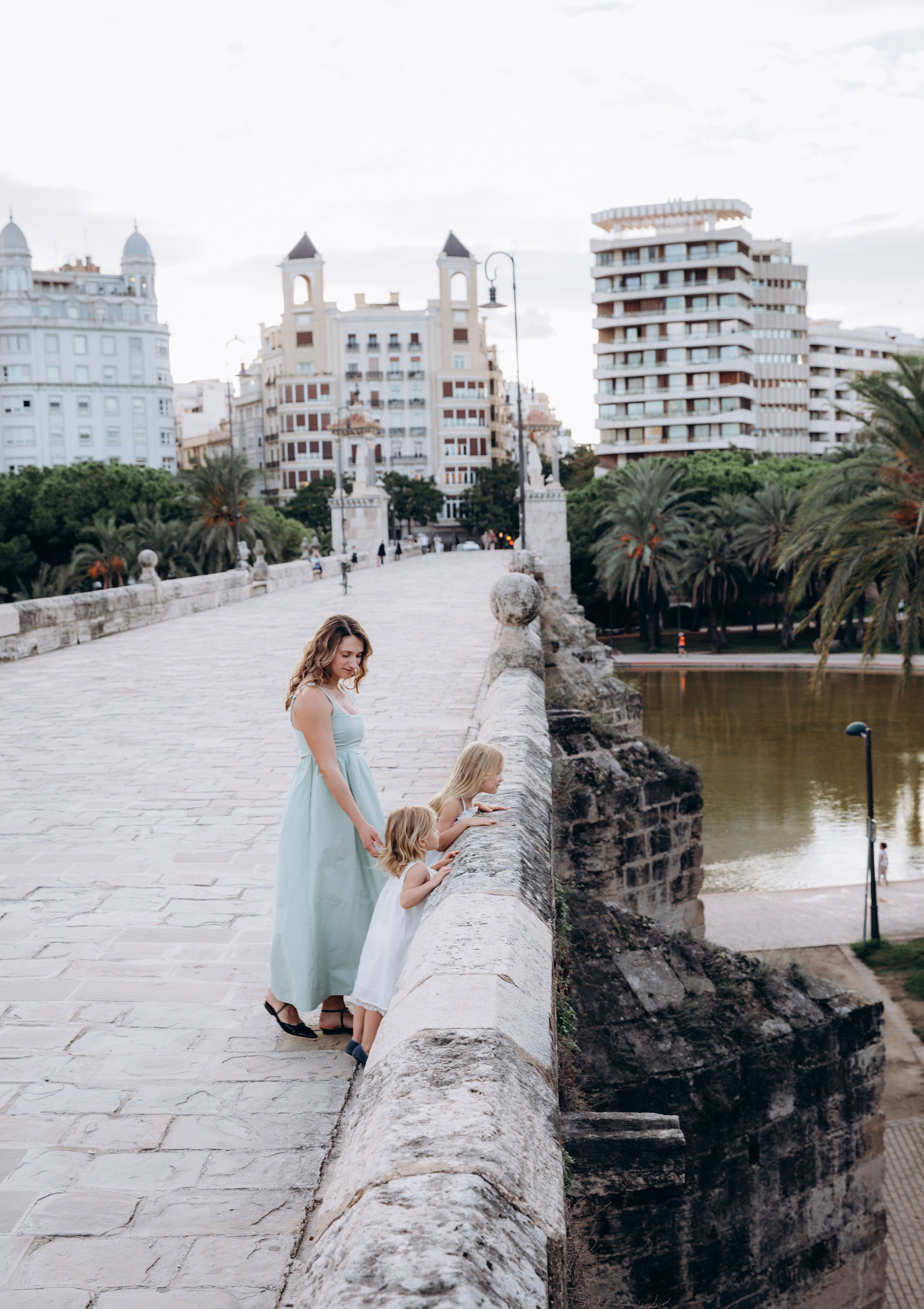 Sesión de fotos familiar espontánea en un puente histórico de Valencia, España — una madre y sus dos hijas disfrutan de un momento de paz con vistas a los Jardines del Turia. Perfecto para quienes buscan fotografía familiar natural y emotiva en Valencia y en toda España, con paisajes urbanos icónicos y encanto arquitectónico.