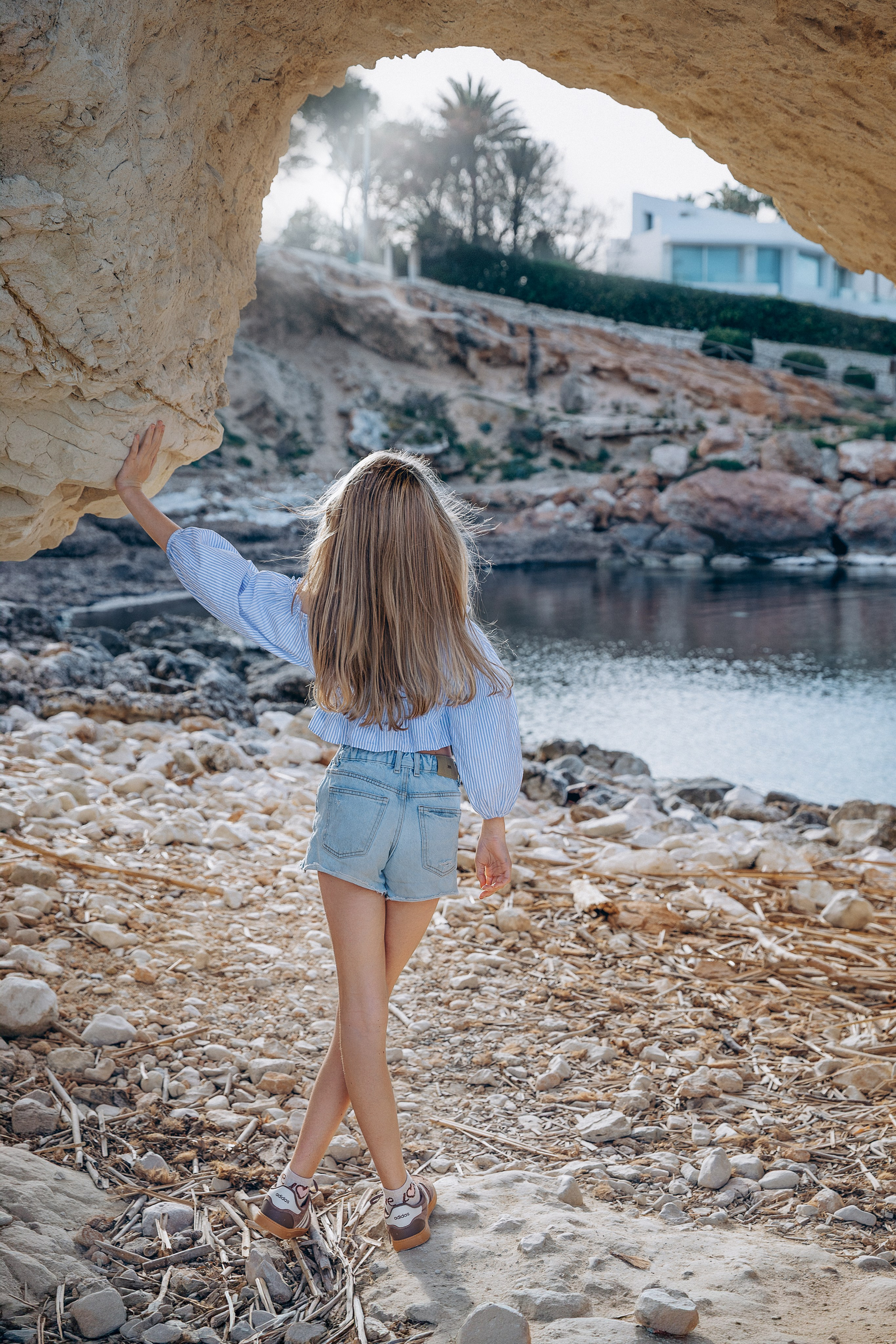 Sesión de retrato femenino en Valencia, España, con una mujer vestida de blanco caminando hacia un arco natural de roca junto al mar. Capturada con suave luz dorada, paisajes costeros y texturas mediterráneas, este retrato artístico lifestyle destaca la libertad, la elegancia y la conexión con la naturaleza, ideal para fotografía de retrato femenino, sesiones individuales, retratos de viaje, sesiones inspiradas en la moda y fotografía al aire libre en Valencia y toda España.