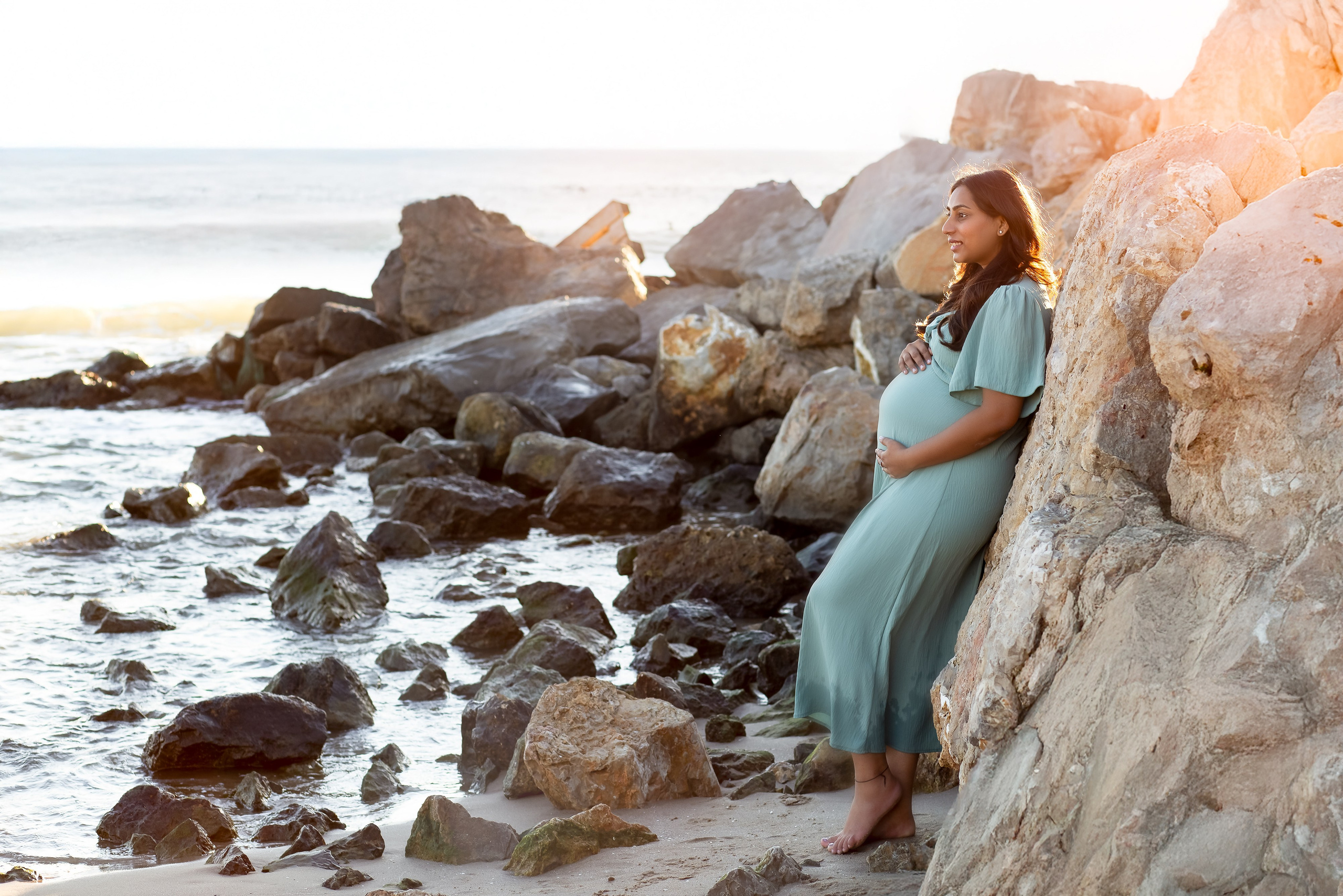 Sesión de fotos de maternidad serena en Valencia, España — una mujer embarazada con un vestido azul suave se recuesta tranquilamente sobre las rocas costeras al atardecer, sosteniendo su barriga. Un momento perfecto para quienes buscan sesiones de embarazo naturales y emotivas en Valencia y en toda España.