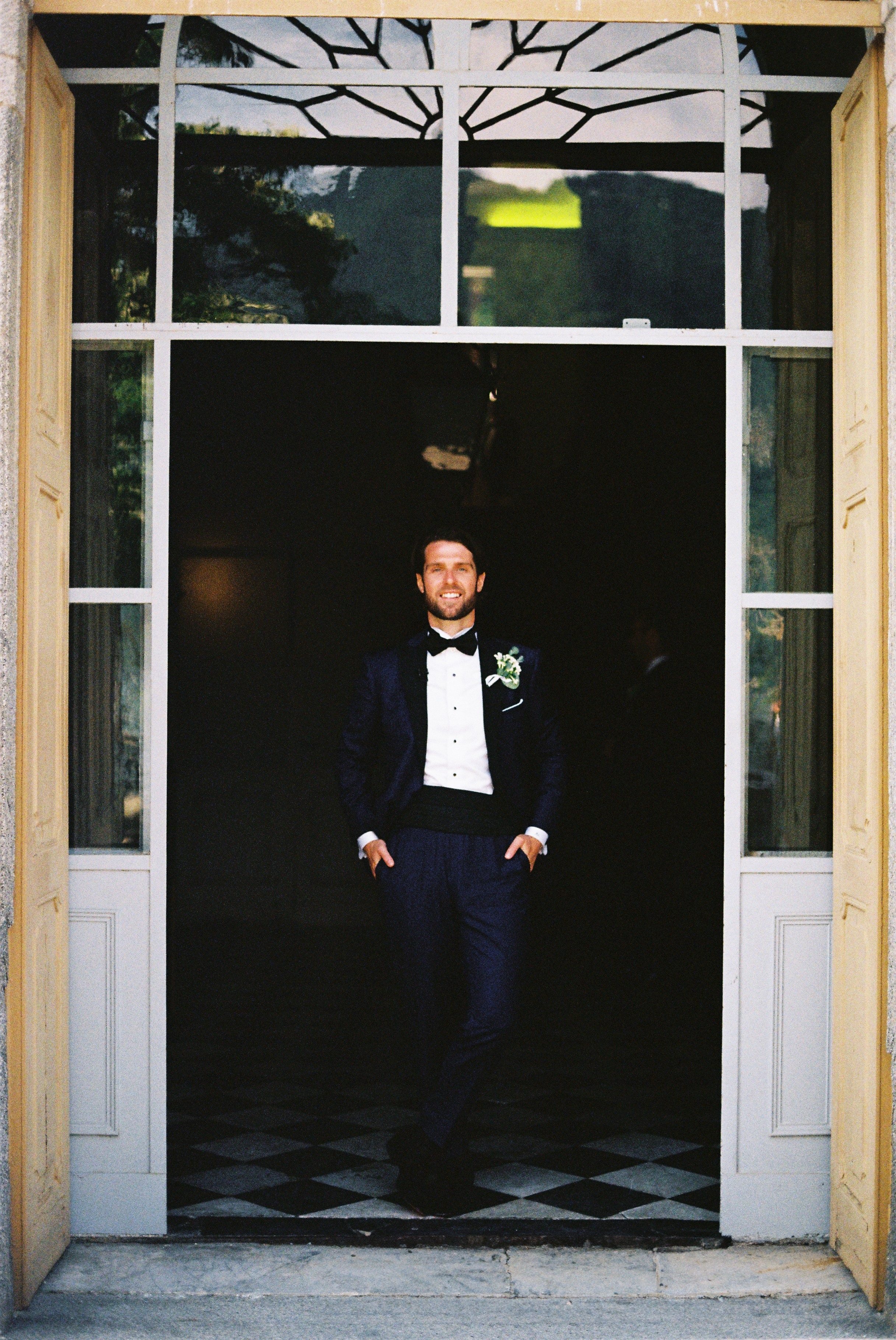Groom in tuxedo smiles, standing in a grand doorway with checkered marble flooring.