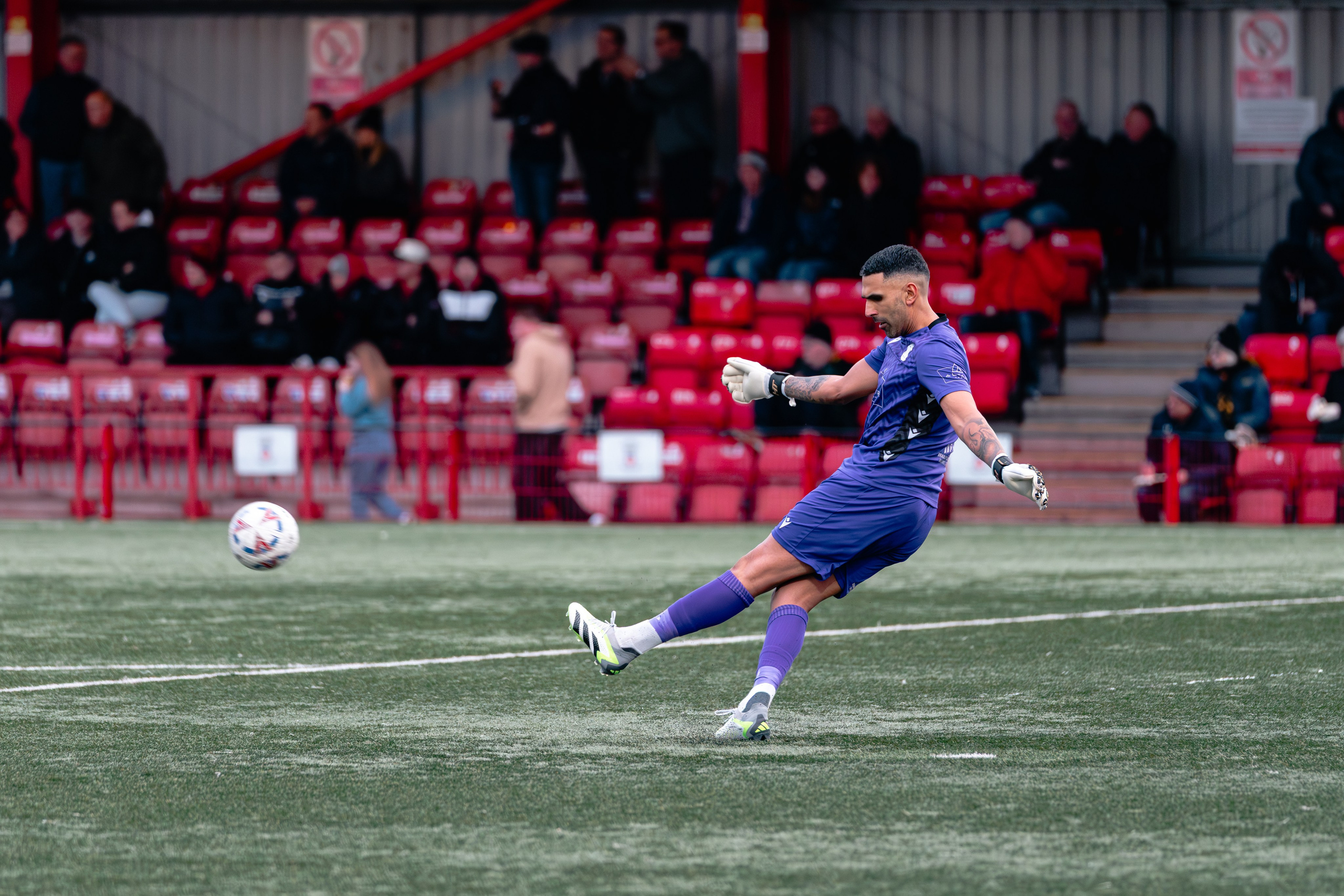 Tamworth goalkeeper Jas Singh in a purple kit plays a long kick upfield during open play.