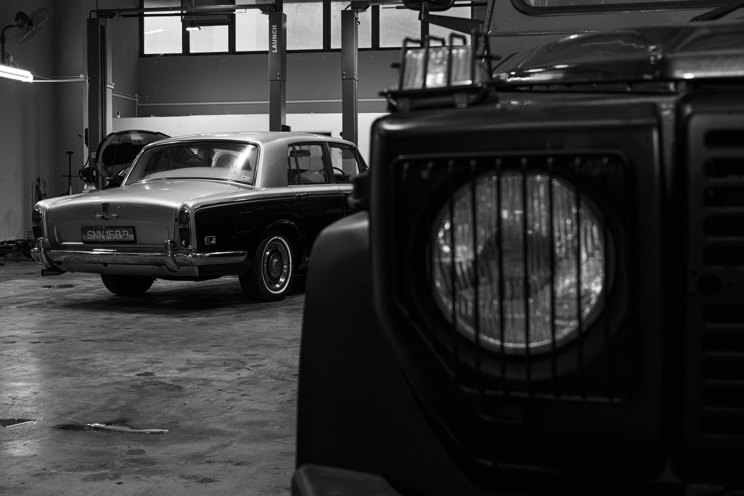 Vintage Rolls-Royce and Mercedes G-Class captured in black-and-white at The Garage by Classic Motorworks in Singapore. This image by MirrorKlezz Photography contrasts the luxury of the Rolls-Royce with the rugged presence of the Mercedes G-Class, showcasing the diversity of classic automotive design within an authentic workshop setting. https://www.classicmotorworks.com.sg/
