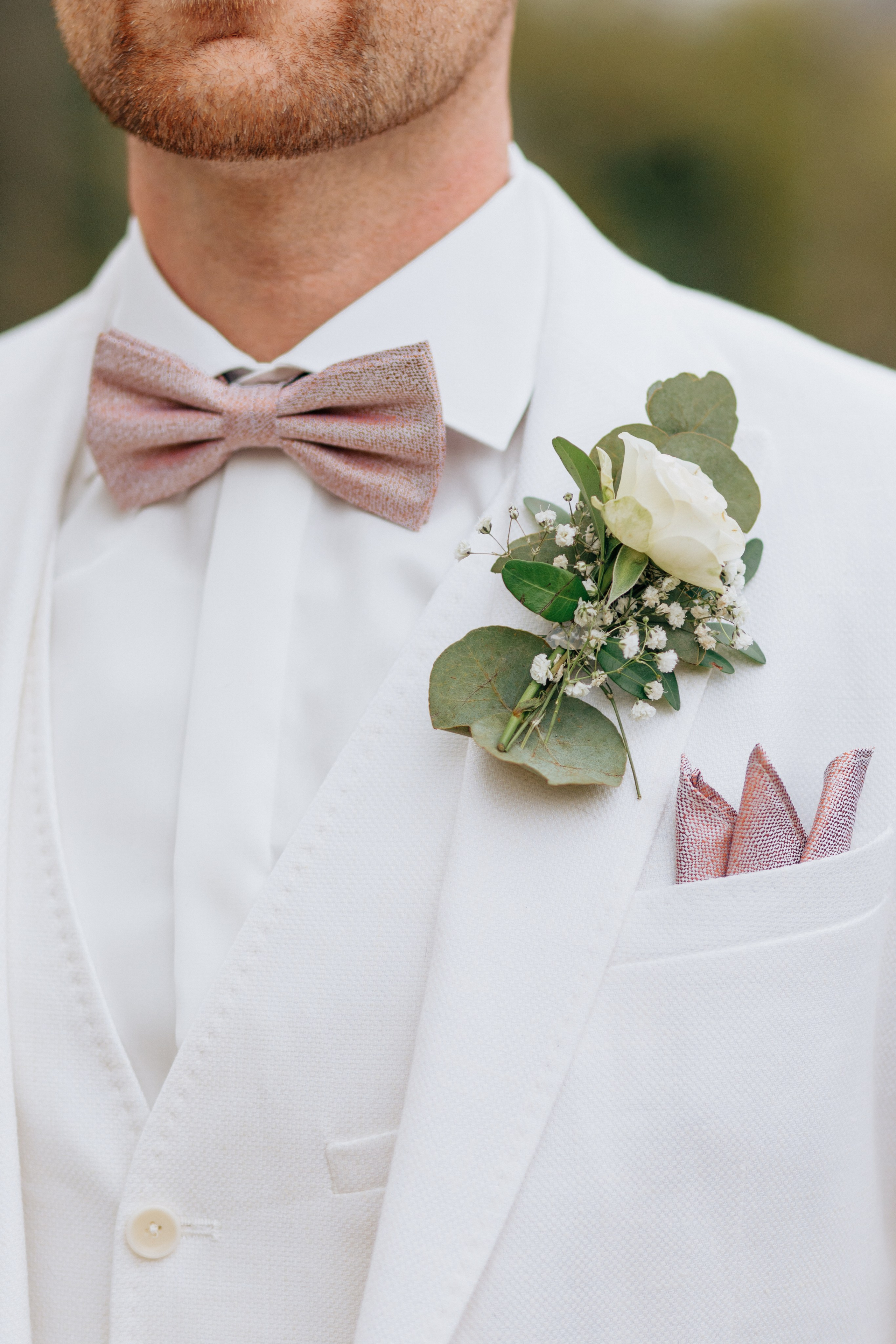 Elegantes Outfit des Bräutigams: Weißer Anzug, rosa Fliege und eine An­steck­blume mit Eukalyptus.