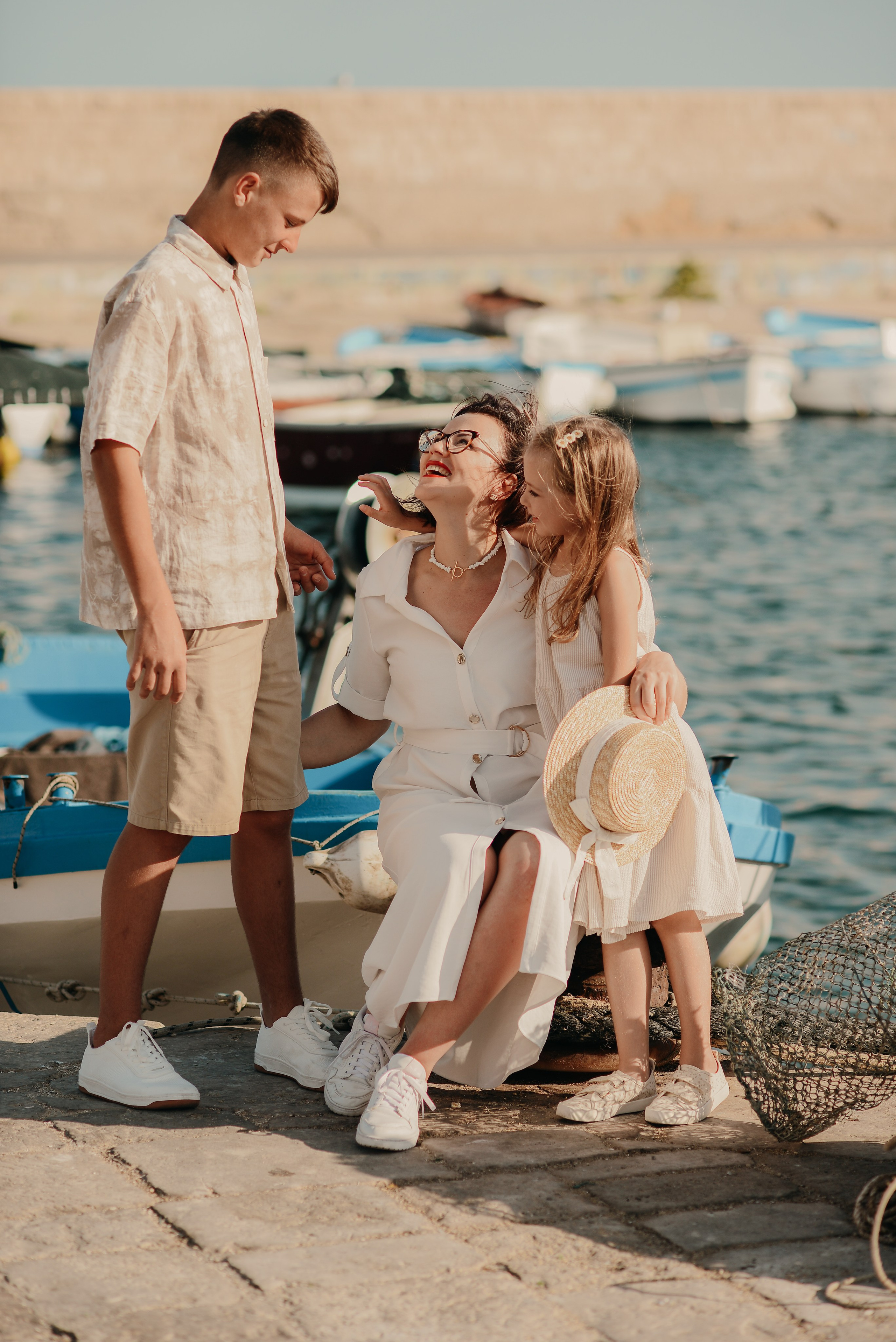 A mother smiling and playing with her children on a sunny day by the harbor of Gallipoli, with boats and the sea in the background.