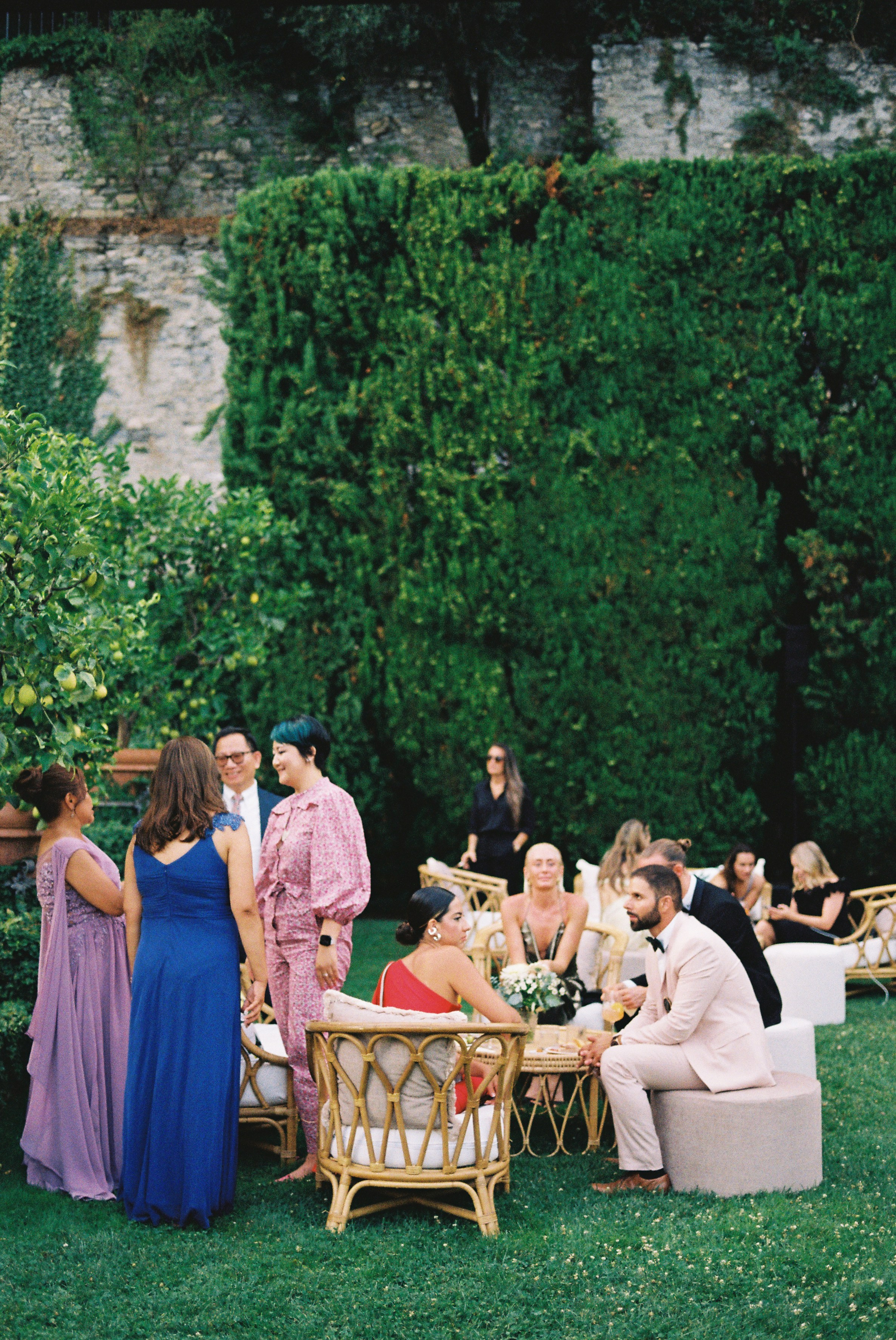 Wedding guests socializing in garden lounge area with greenery and casual seating.