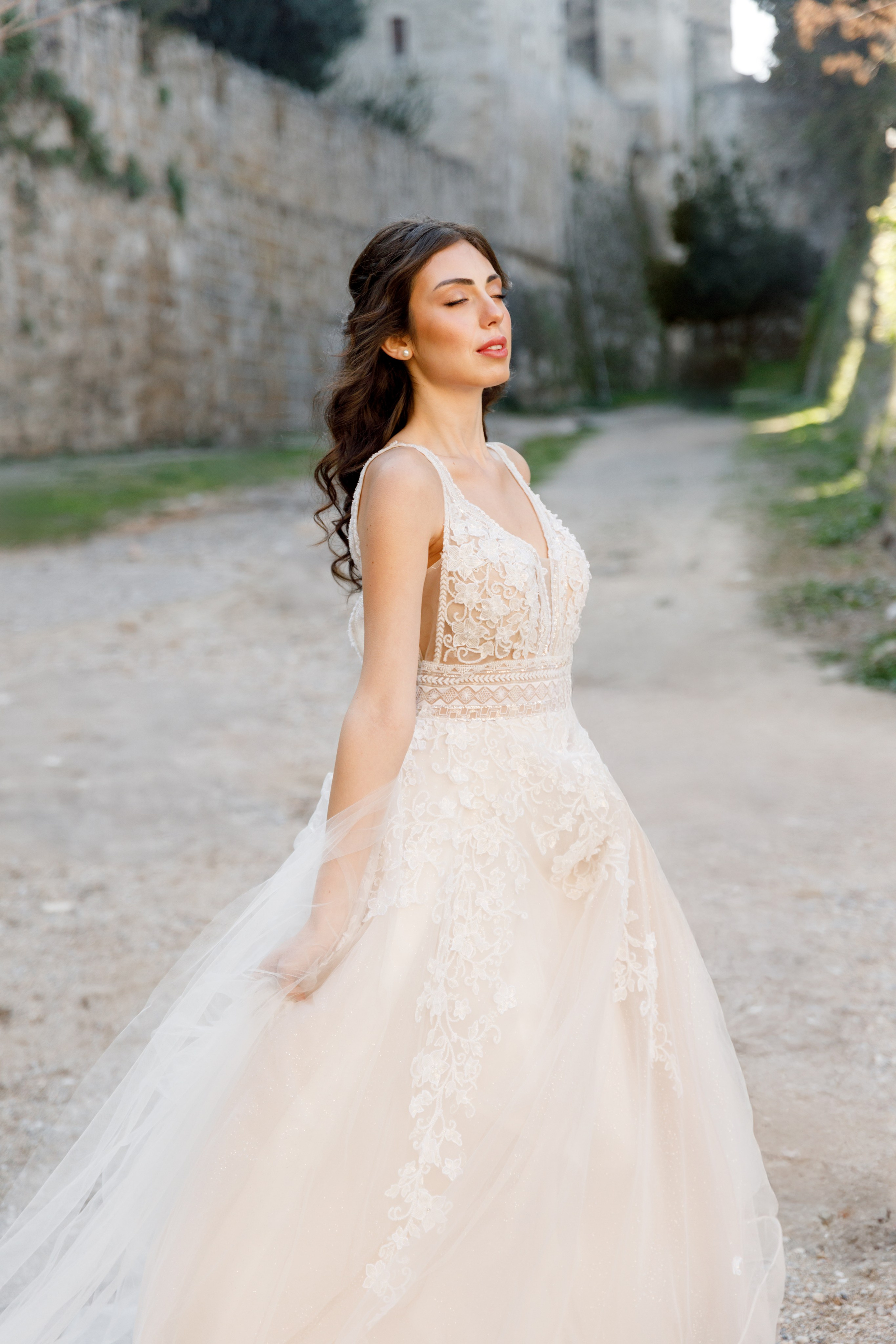 A stunning bride gazes thoughtfully in the enchanting alleys of Rhodes' Old Town, her flowing wedding dress complementing the rustic charm of the cobblestone streets and ancient architecture. The editorial-style portrait captures her poise and the romantic atmosphere of the medieval surroundings, bathed in warm, golden light.