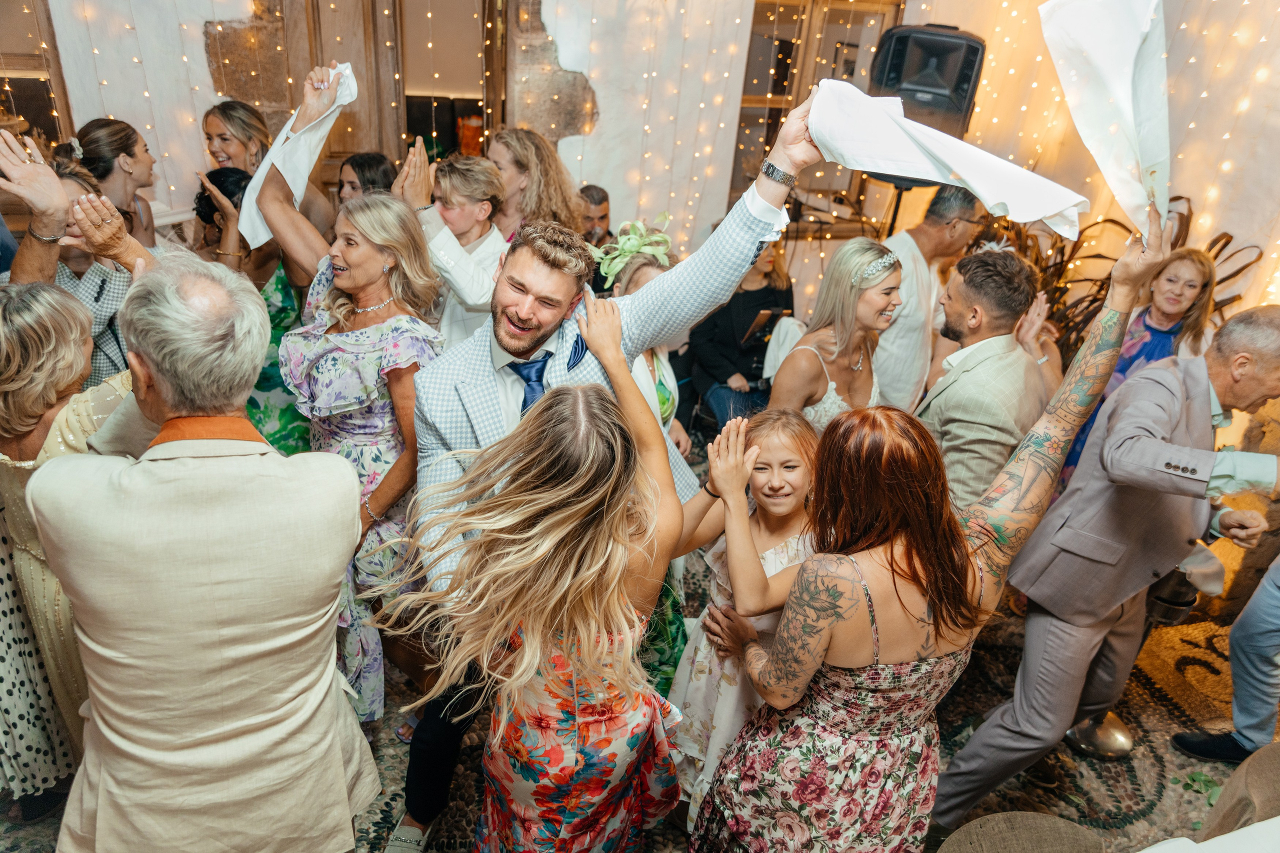 Guests waving white handkerchiefs in the air during a lively traditional dance at an English-style wedding party held in a picturesque location in Greece, celebrating the newlyweds with joy and enthusiasm.