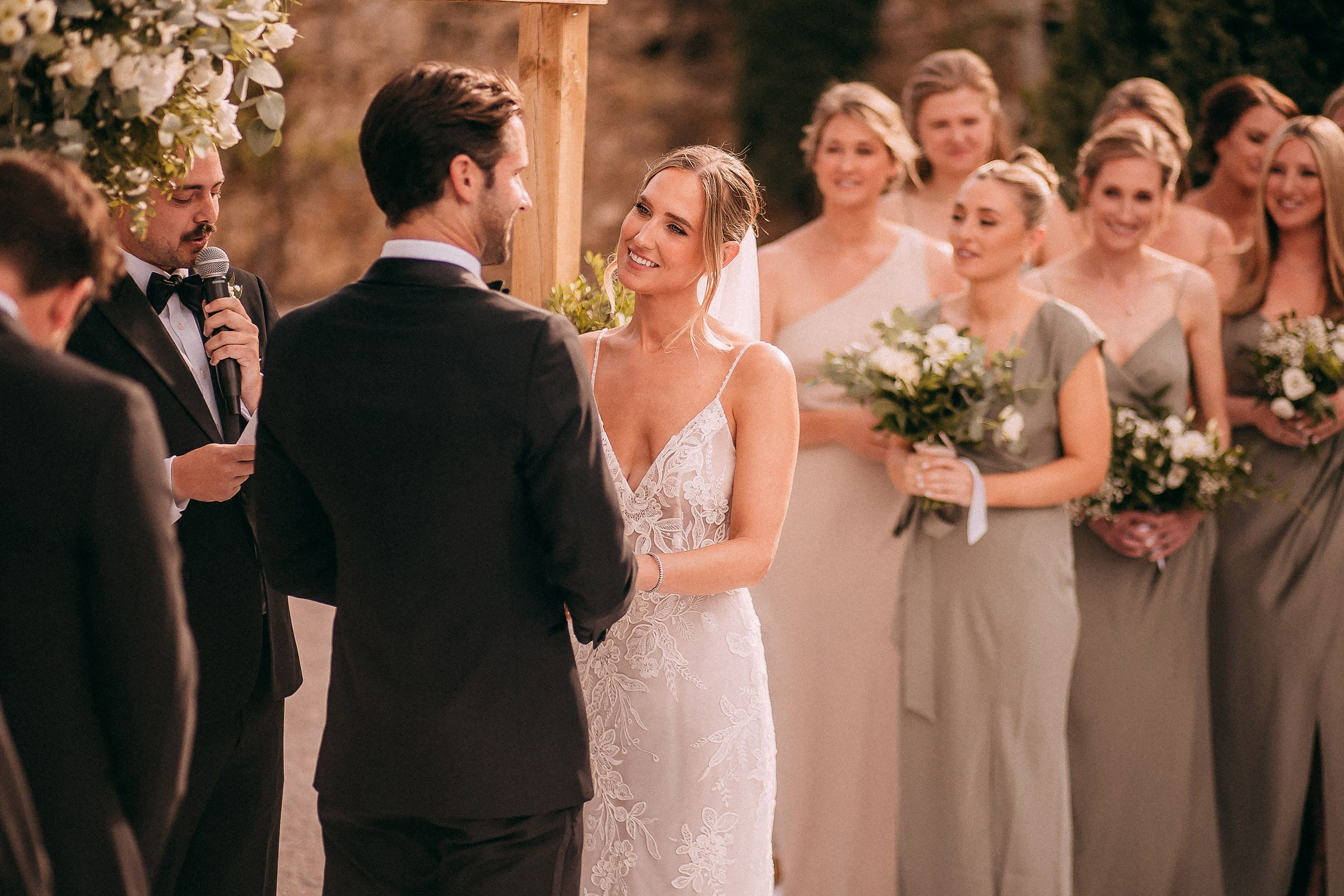 The bride and groom facing each other under the wedding arch, exchanging smiles as the officiant speaks during their vows.