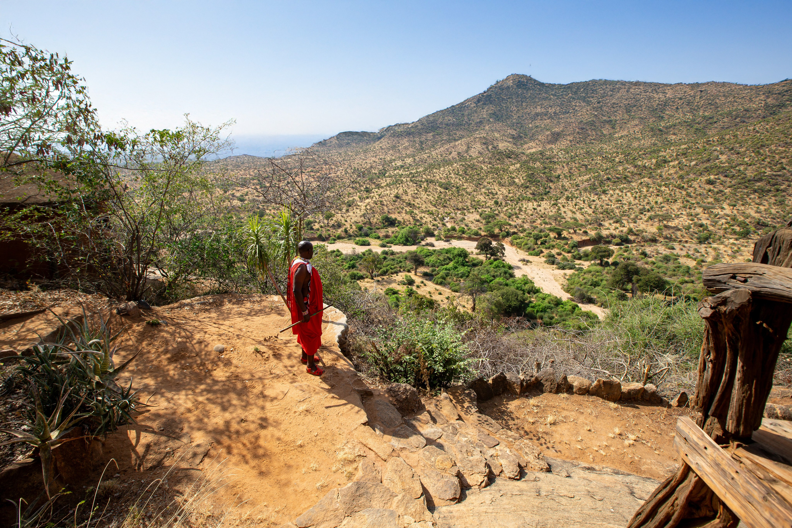 A wide angle photo of vast land and hills in the background. A Maasai elder is standing in the foreground overlooking the vastness. Documentary storytelling