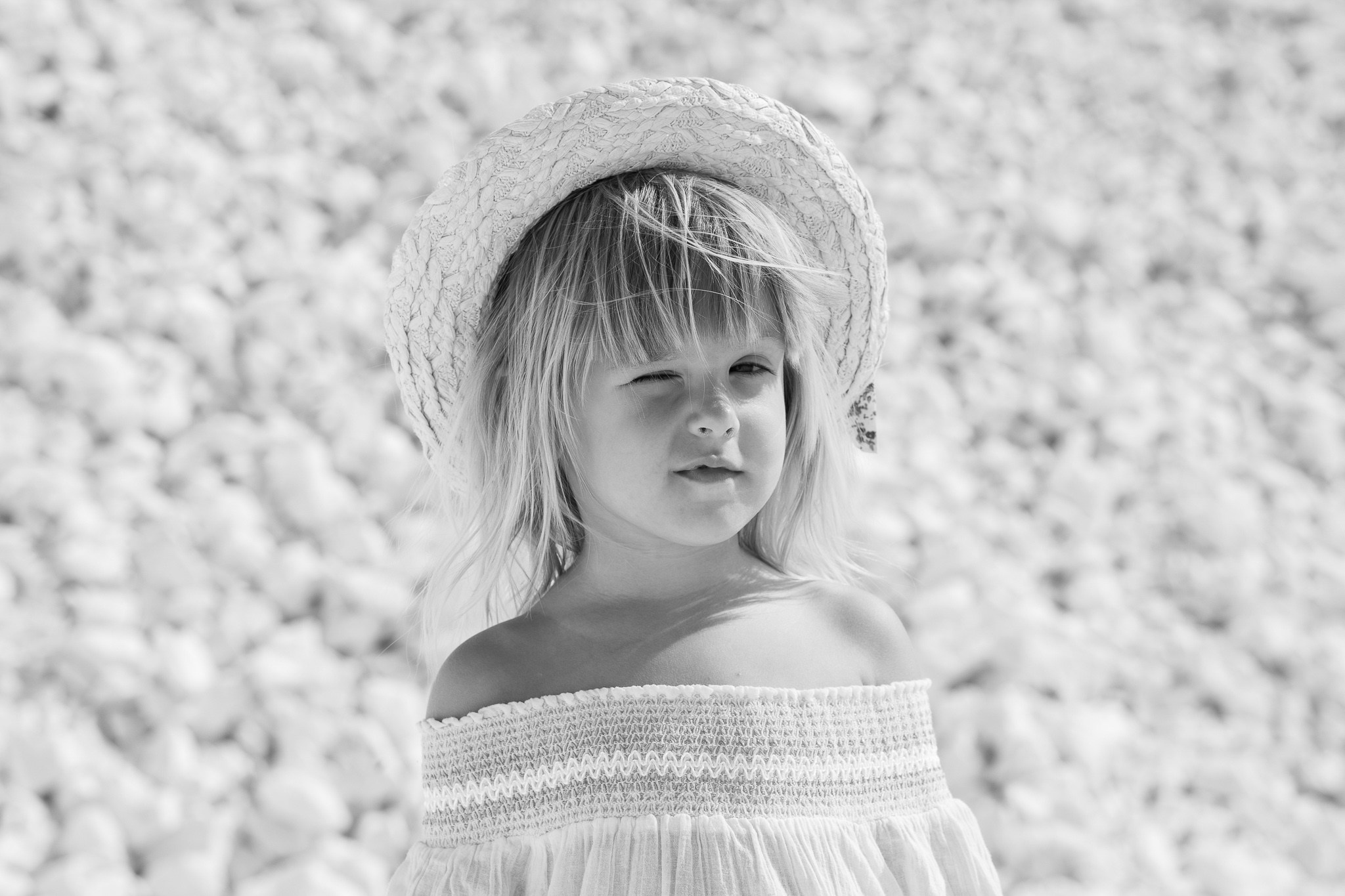 Crno-bela fotografija razigrane mlade devojčice u slamnatom šeširu i topu spuštenih ramena, koja namiguje kameri na šljunkovitoj plaži.