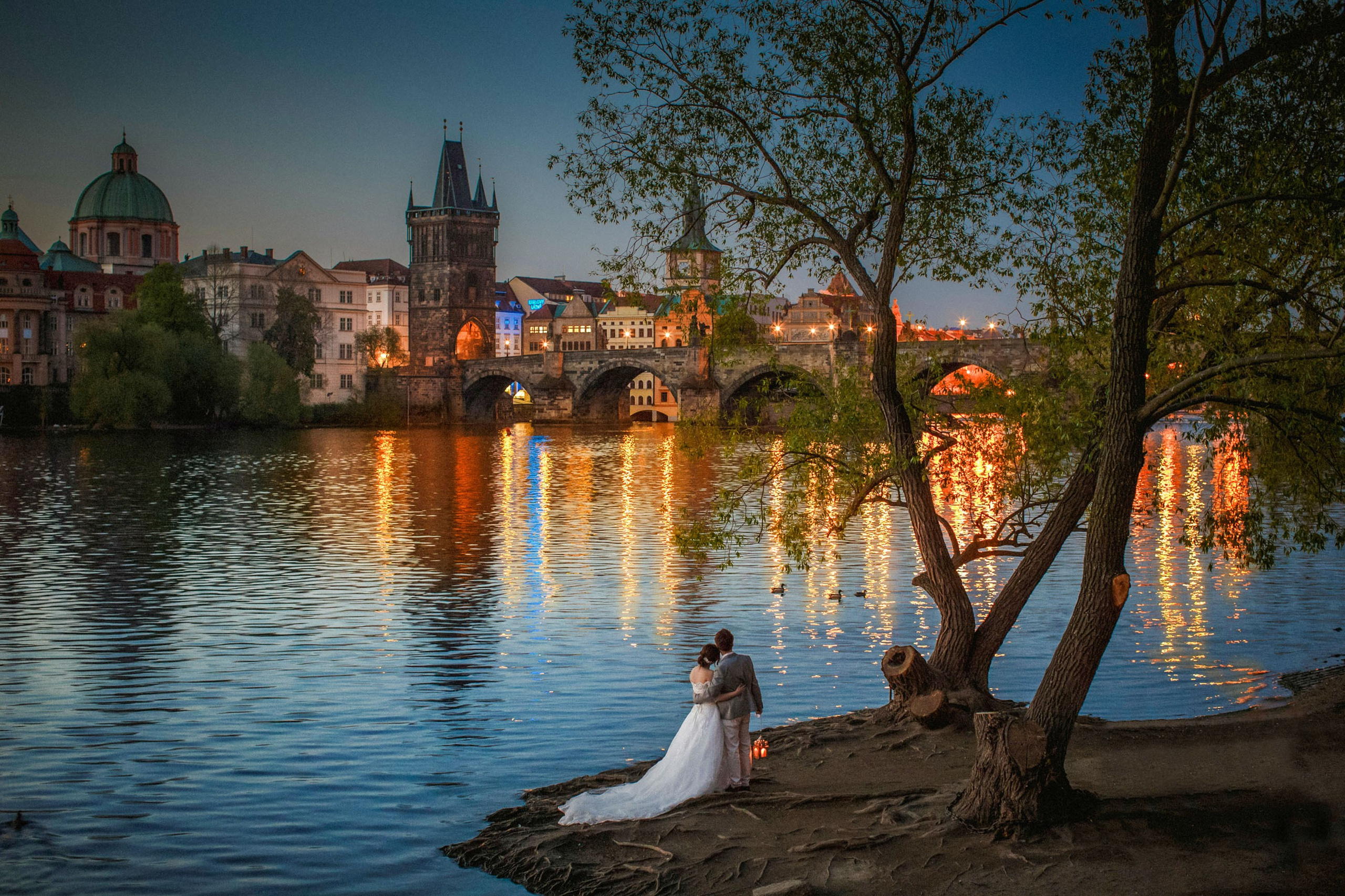 Newlyweds sharing a lantern-lit moment, looking towards the beautifully illuminated Charles Bridge with its historic gas lamps in nighttime Prague.