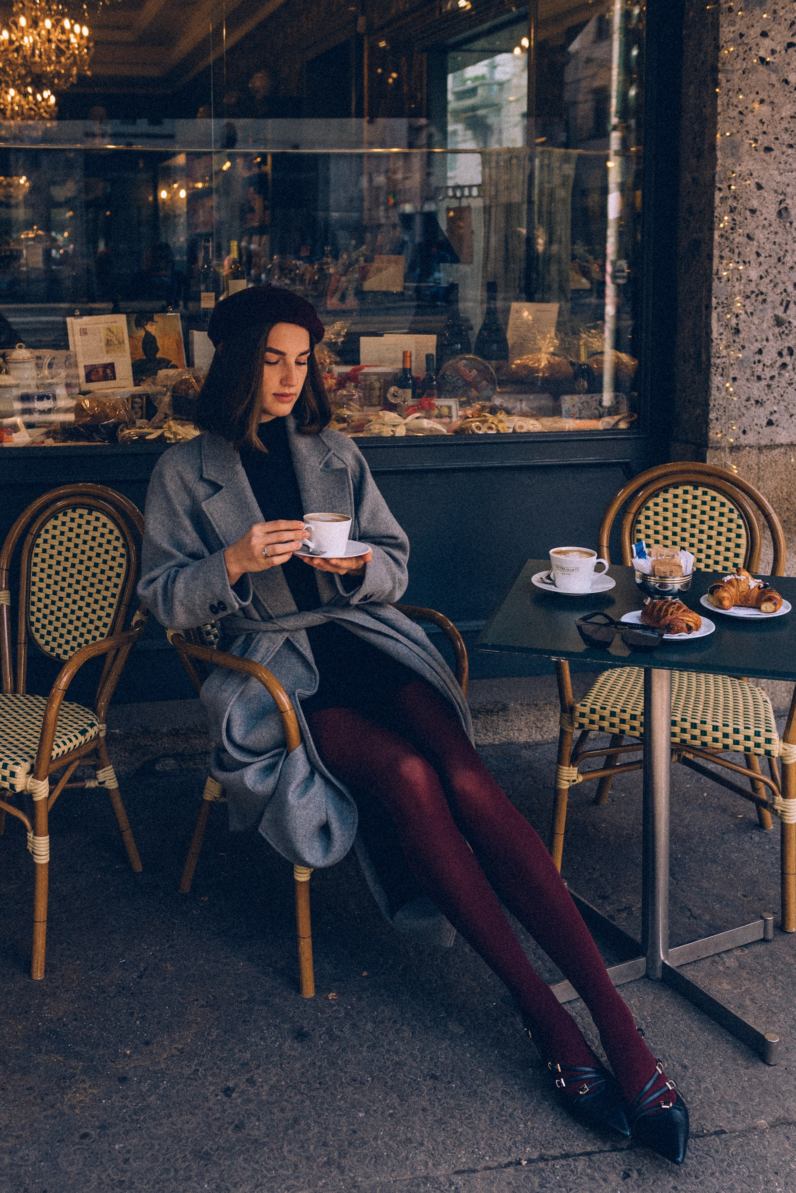 Elegant woman sitting at a Milanese cafe, enjoying a cappuccino paired with fresh pastries, dressed in a gray coat and burgundy tights