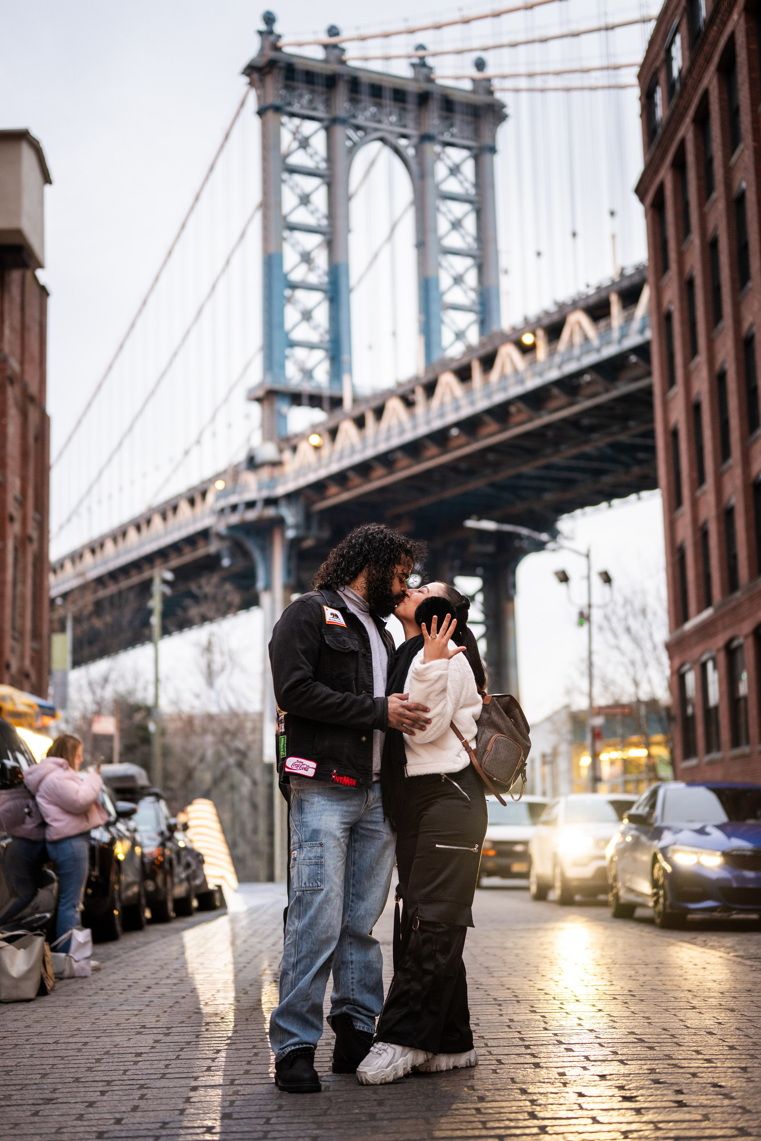 Wide shot of couple standing in the street in DUMBO with Manhattan Bridge view, iconic Brooklyn photo location.