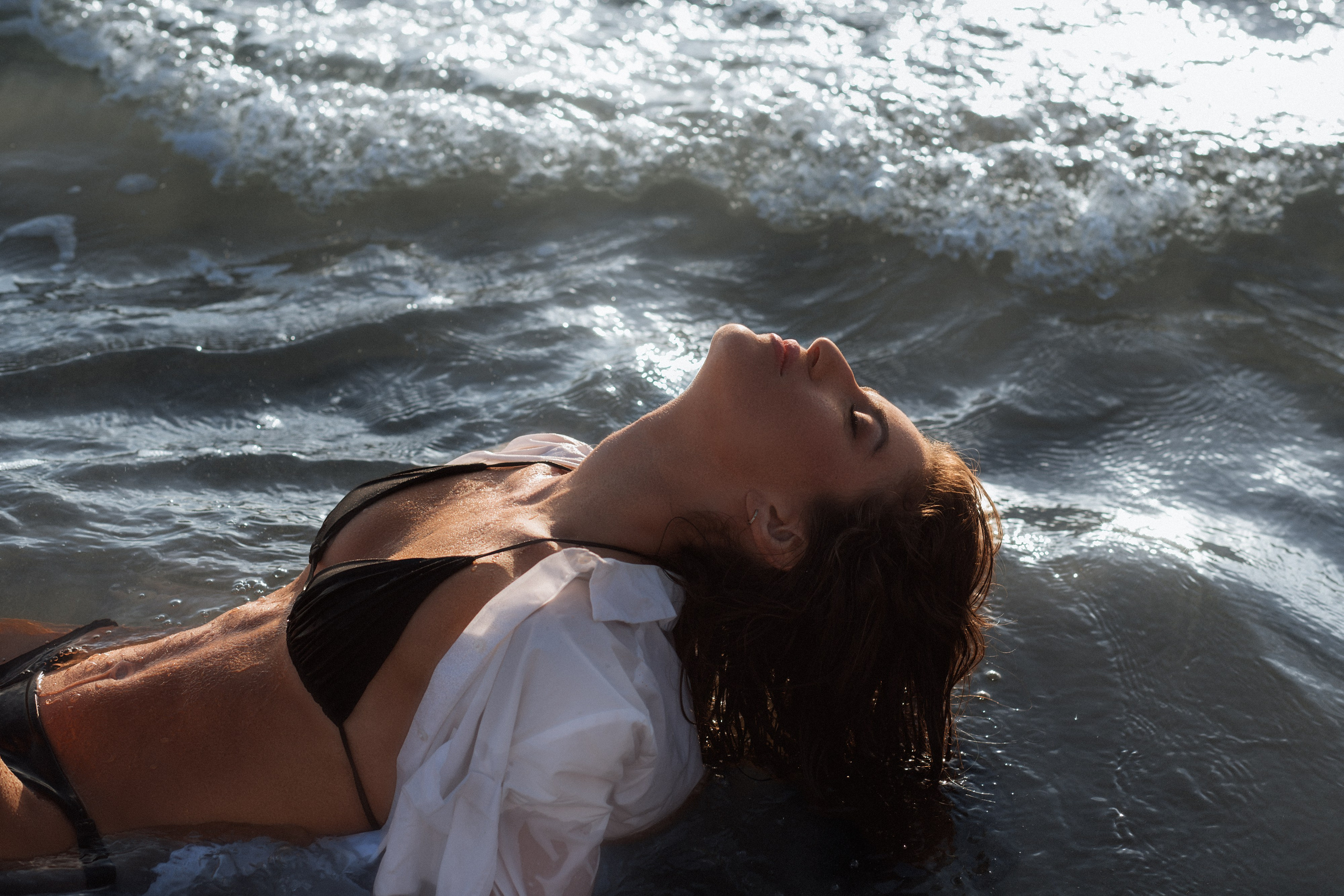 Чувственная пляжная фотосессия в Валенсии, Испания — девушка в чёрном бикини и белой рубашке отдыхает в море на закате, излучая уверенность, элегантность и природную красоту. Идеальный выбор для тех, кто ищет художественную и вдохновляющую портретную съёмку в Валенсии и на побережье Испании.