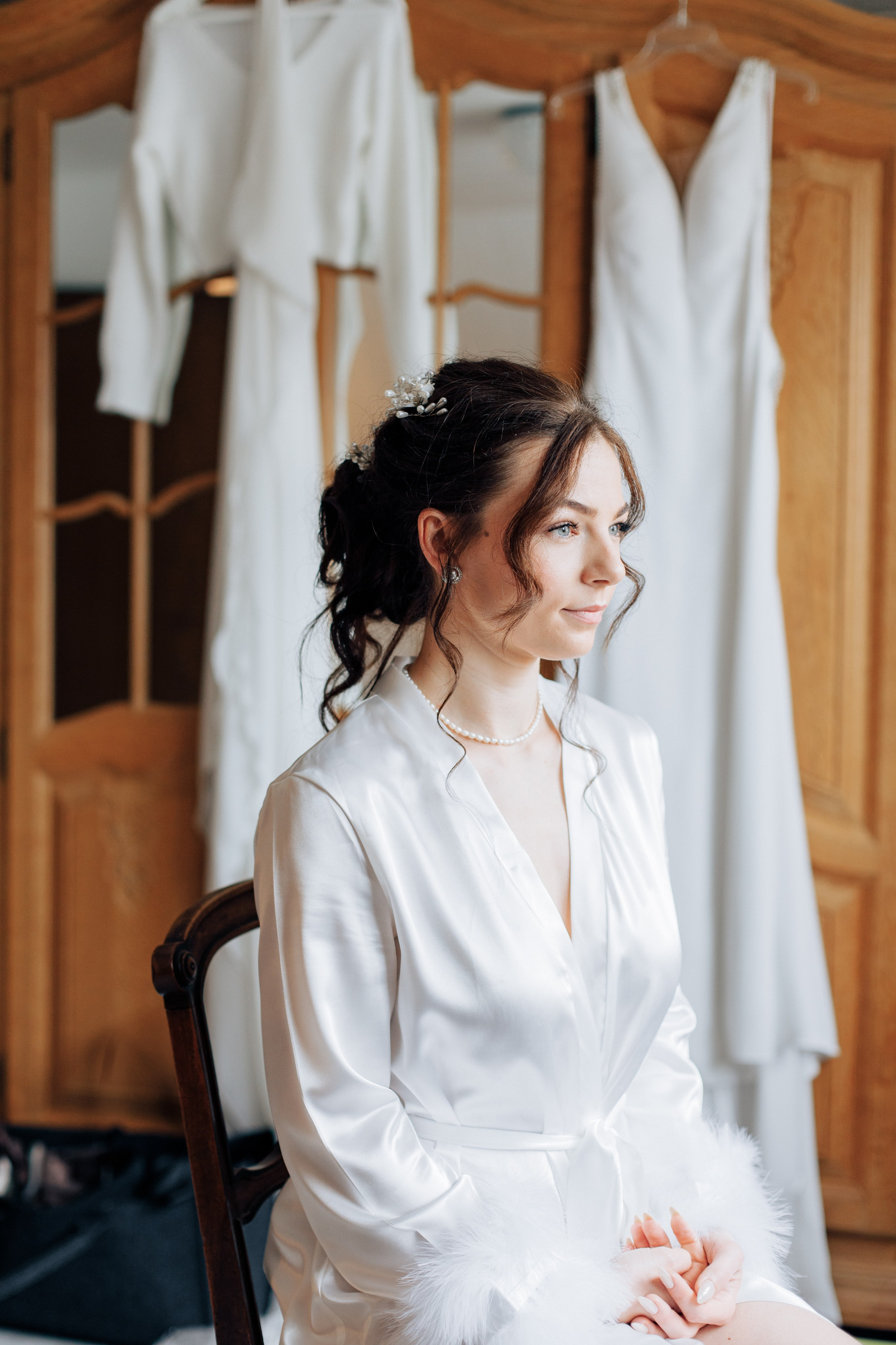 Die Braut sitzt nachdenklich auf einem Stuhl und blickt aus dem Fenster, während ihr Hochzeitskleid im Hintergrund an einem Holzschrank hängt.