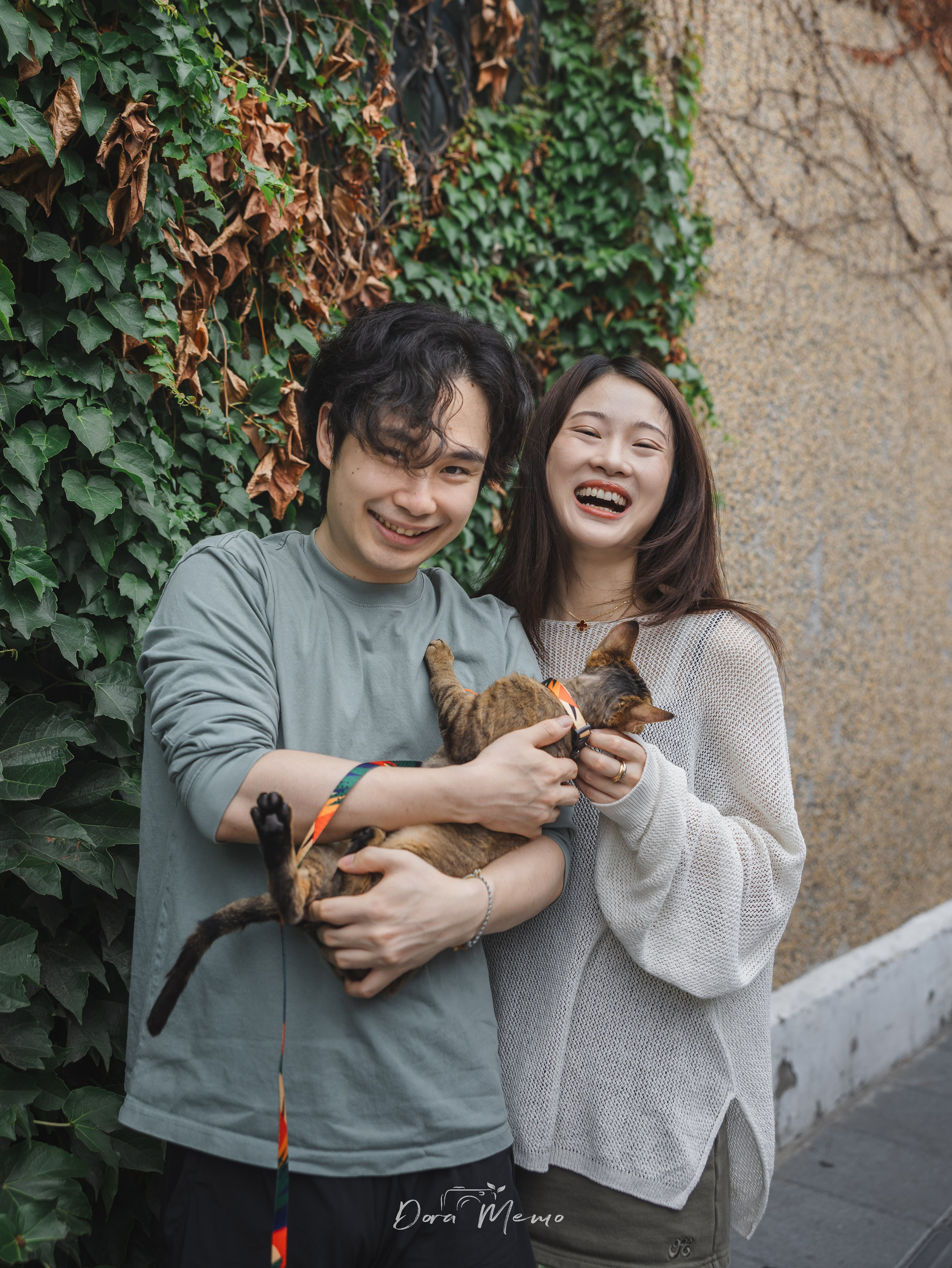  A couple holds their cat outside near an ivy-covered wall during a natural pet family photo session.