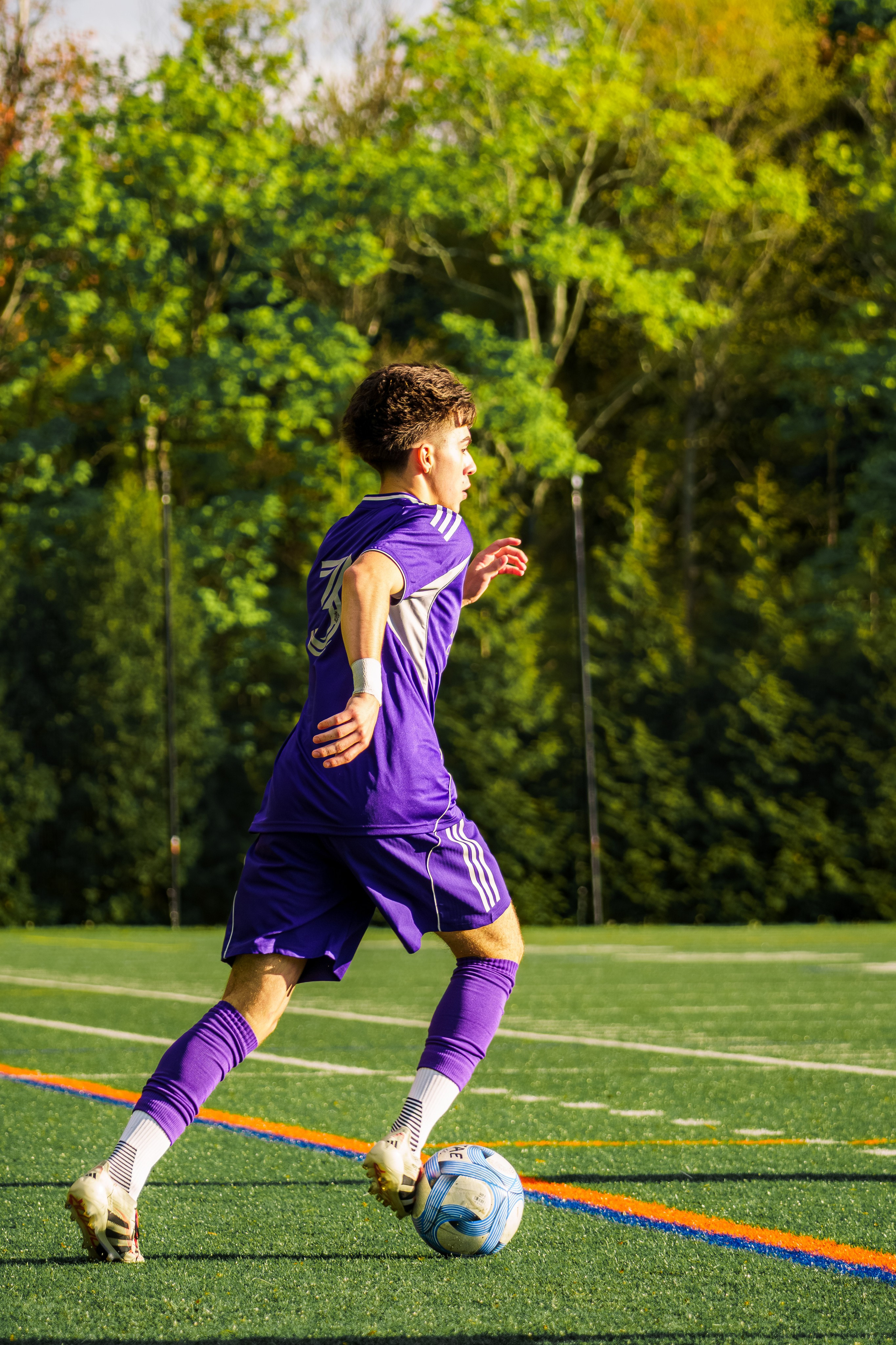 A soccer player in a purple uniform is moving along the sideline during an outdoor match. Sprinting with the ball on his feet, creating an attack. 