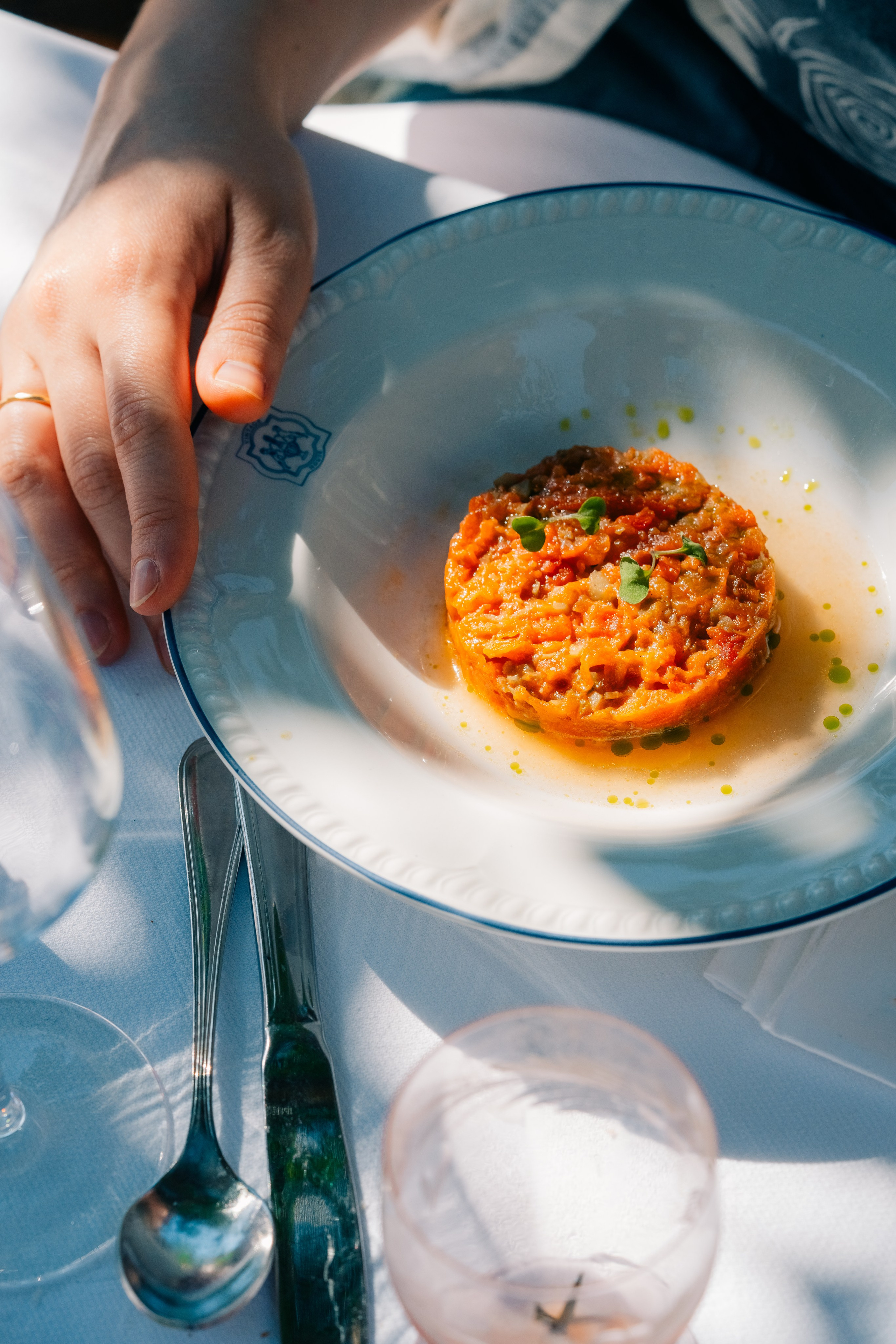 Рука тянется к тарелке изысканного блюда, поданного на открытом воздухе. Рука тянется к изысканному блюду из мелко нарезанных овощей, поданному на белой тарелке, на открытом воздухе на залитом солнцем столе.