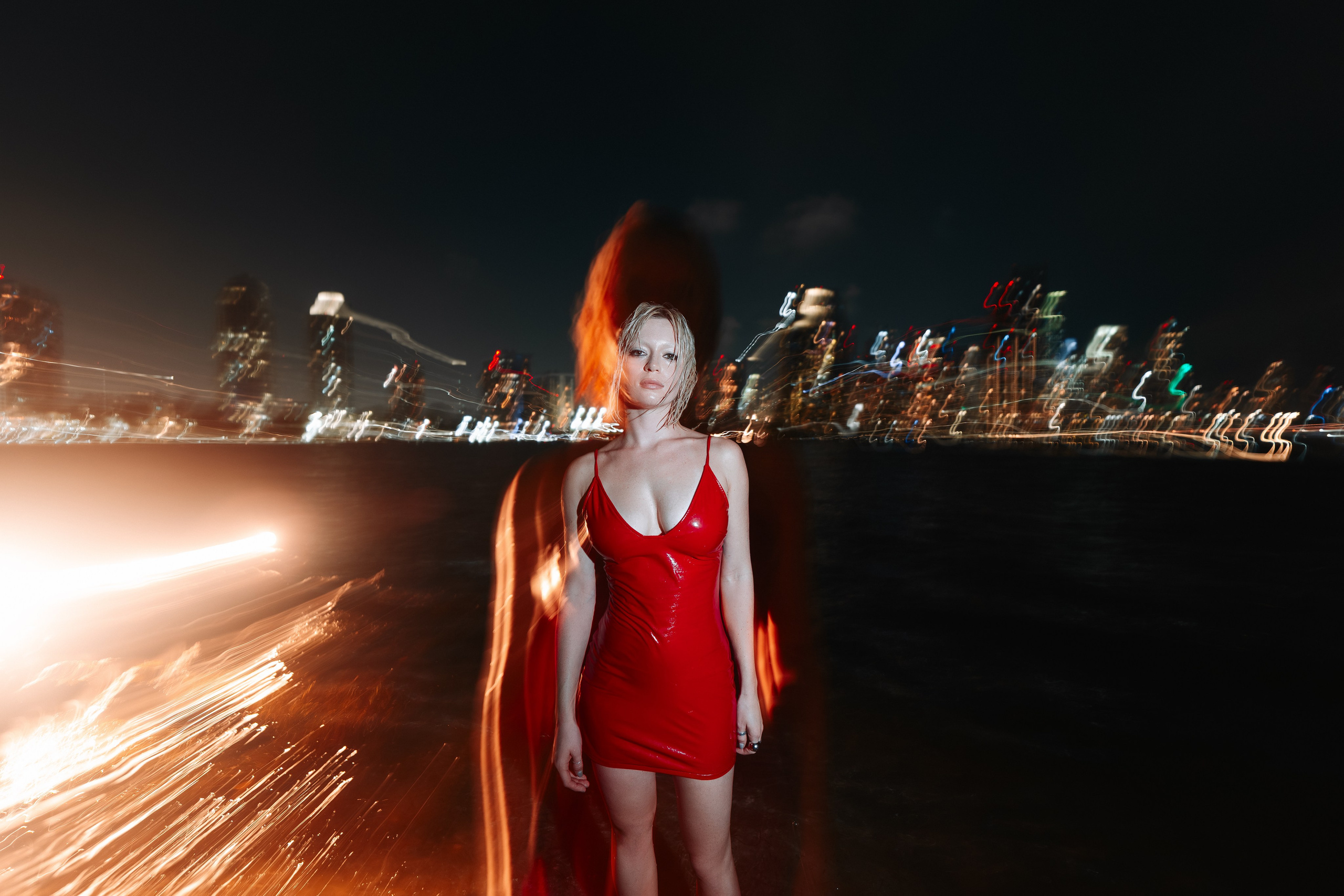 Dynamic shot with a soft motion blur created by long exposure. The model in a red dress walks barefoot along the waterline, Miami lights fading into movement. Professional night shoot with cinematic hard-light shaping. Miami photographer, cinematic portraits, night session.