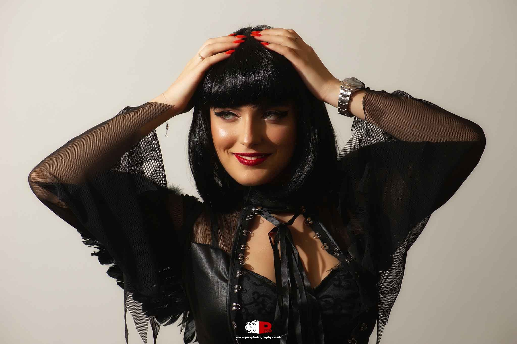 Stylish woman in a gothic-inspired black outfit with red nails posing with hands on her head.