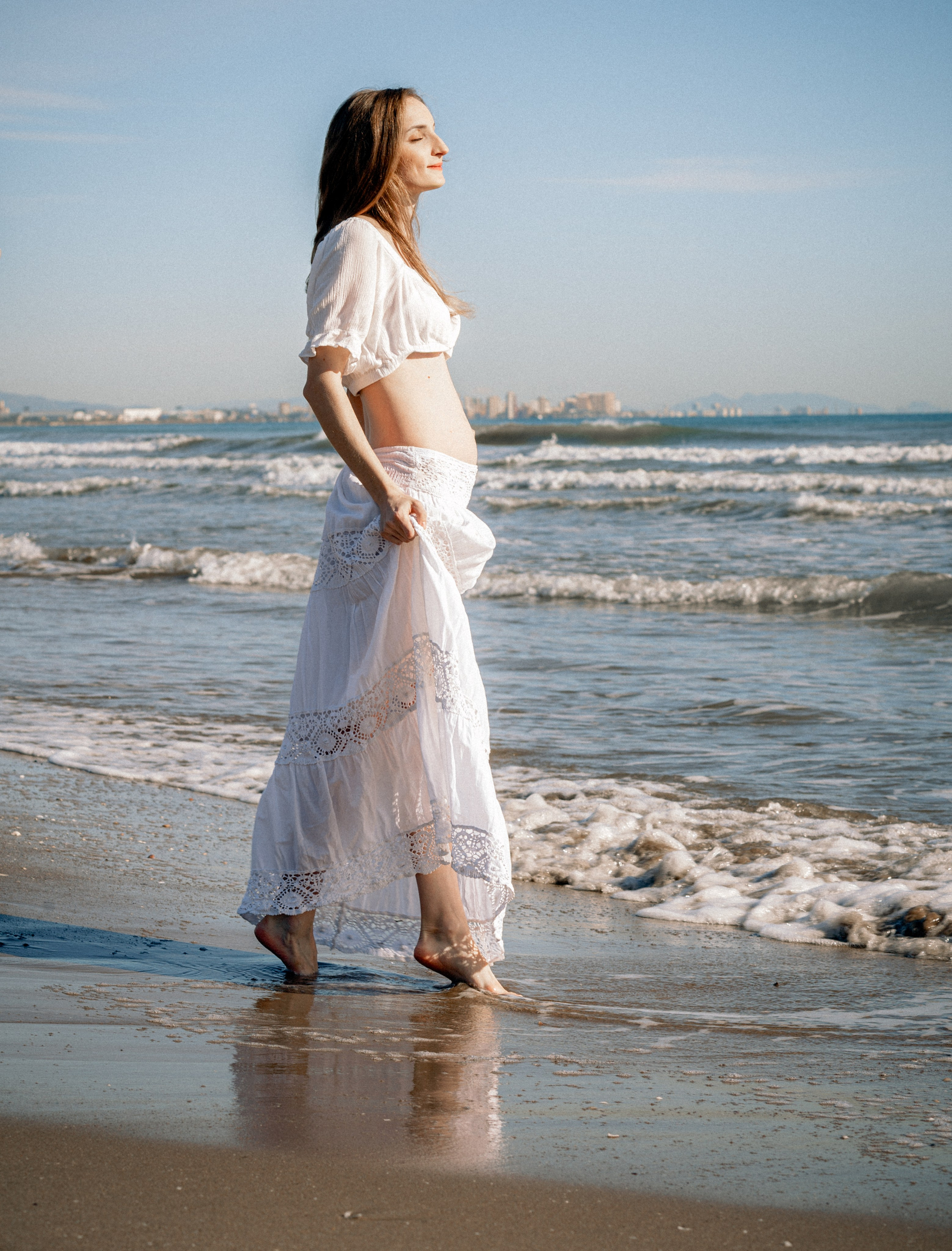 Sesión de fotos de embarazo en València, España, con una mujer embarazada caminando descalza por la playa mediterránea, capturada con luz solar natural suave, olas delicadas, un vestido blanco fluido y una atmósfera tranquila y emocional — ideal para fotografía de embarazo, sesiones de maternity, sesiones de embarazo en la playa, retratos lifestyle de embarazo y sesiones románticas de embarazo en València y en toda España.