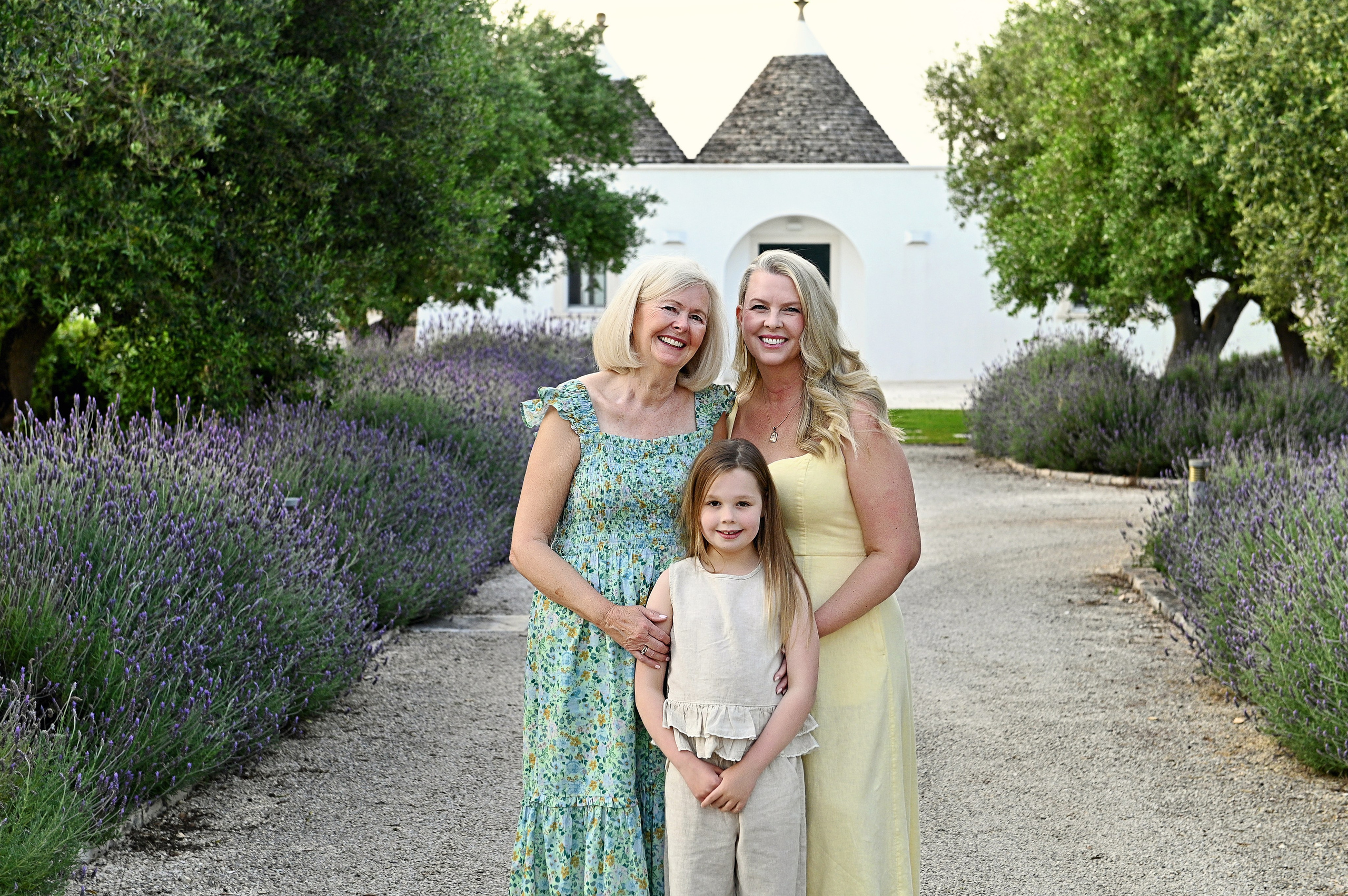 Servizio fotografico di famiglia in Puglia: tre generazioni — nonna, mamma e figlia posano su un sentiero di lavanda con un tradizionale trullo sullo sfondo.