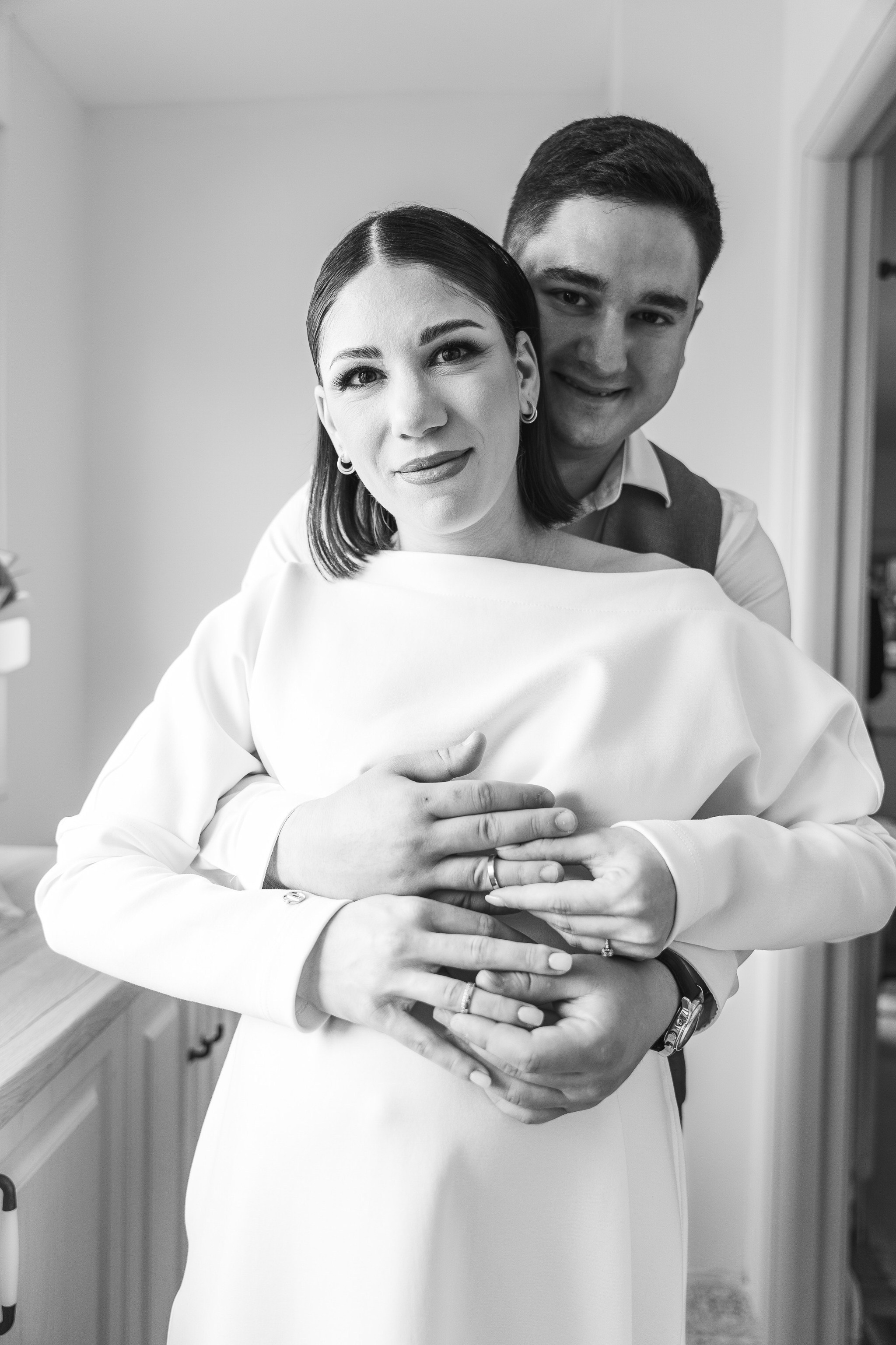 Crno-bela fotografija mladenaca u zatvorenom prostoru, gde mladoženja stoji iza mlade i grli je dok oboje gledaju u kameru, sa rukama nežno preklopljenim preko njenog struka.