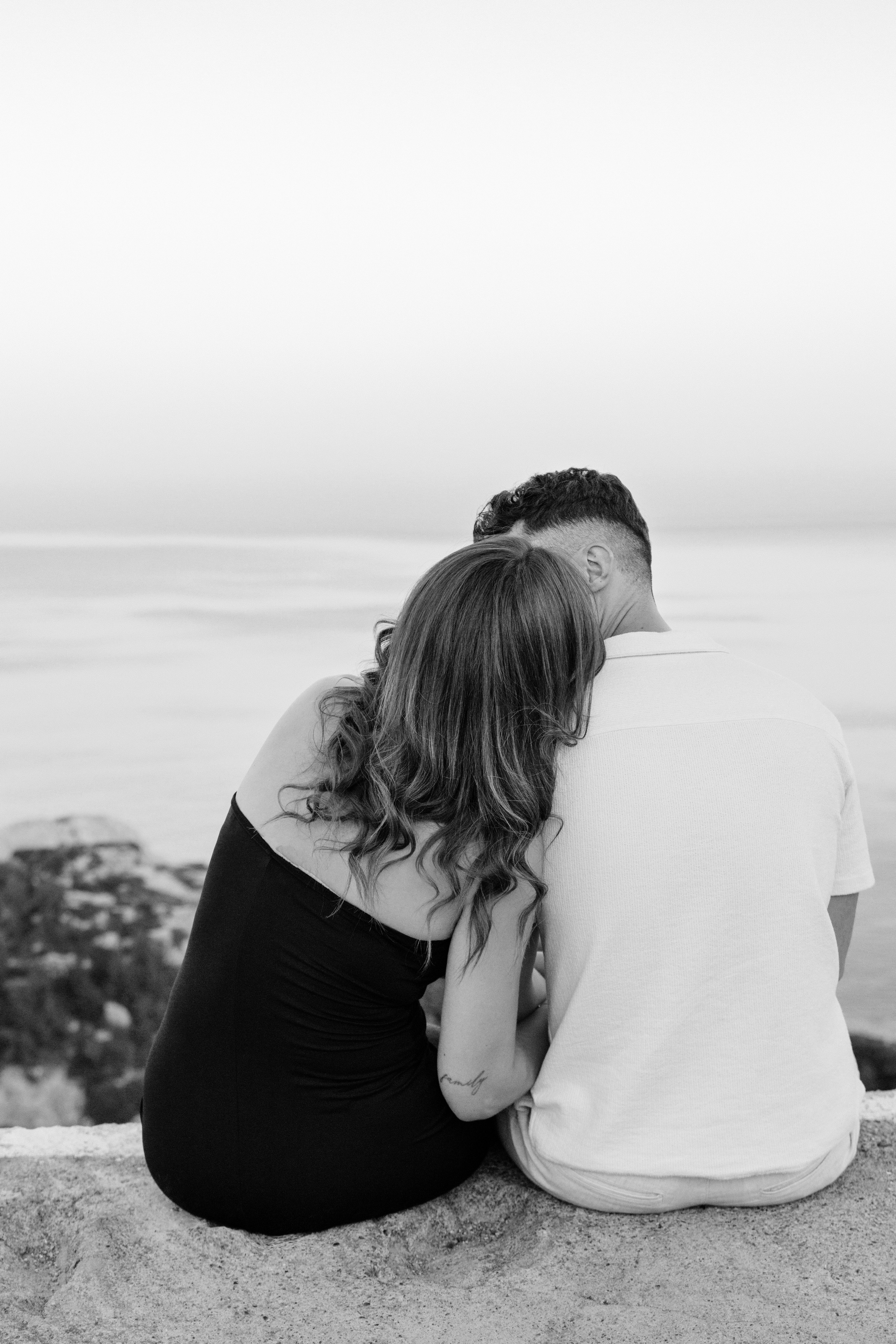 "Rückenansicht eines Paares, das nebeneinander auf den Felsen des Profitis Ilias sitzt. Sie schauen gemeinsam auf das tiefblaue Ägäische Meer und die Küste von Rhodos unter ihnen. Ein Moment des Innehaltens nach dem Heiratsantrag in der friedlichen Natur Griechenlands."