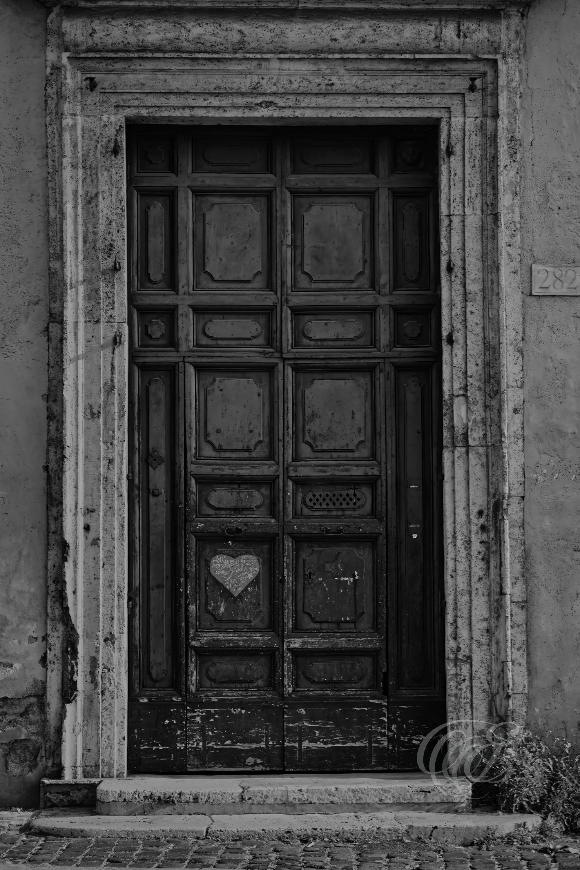 Street Photography — Rome, Italy — Old Door + Heart — B&W — Black and white study of an aged Roman doorway with a small heart marking, emphasizing weathered surfaces, architectural details, and urban texture — Eduardo Bartoli Photography.