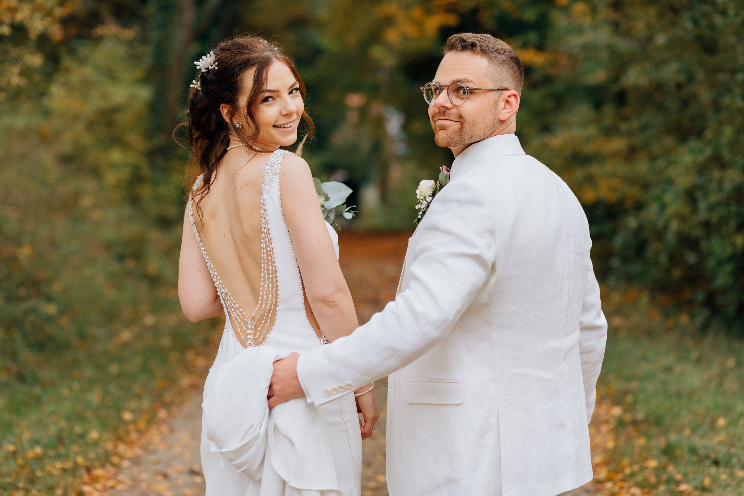 Der Bräutigam und die Braut stehen mit dem Rücken zueinander, drehen sich um, schauen lachend über ihre Schultern in die Kamera, während der Bräutigam den Schleppe ihres Hochzeitskleides hält.