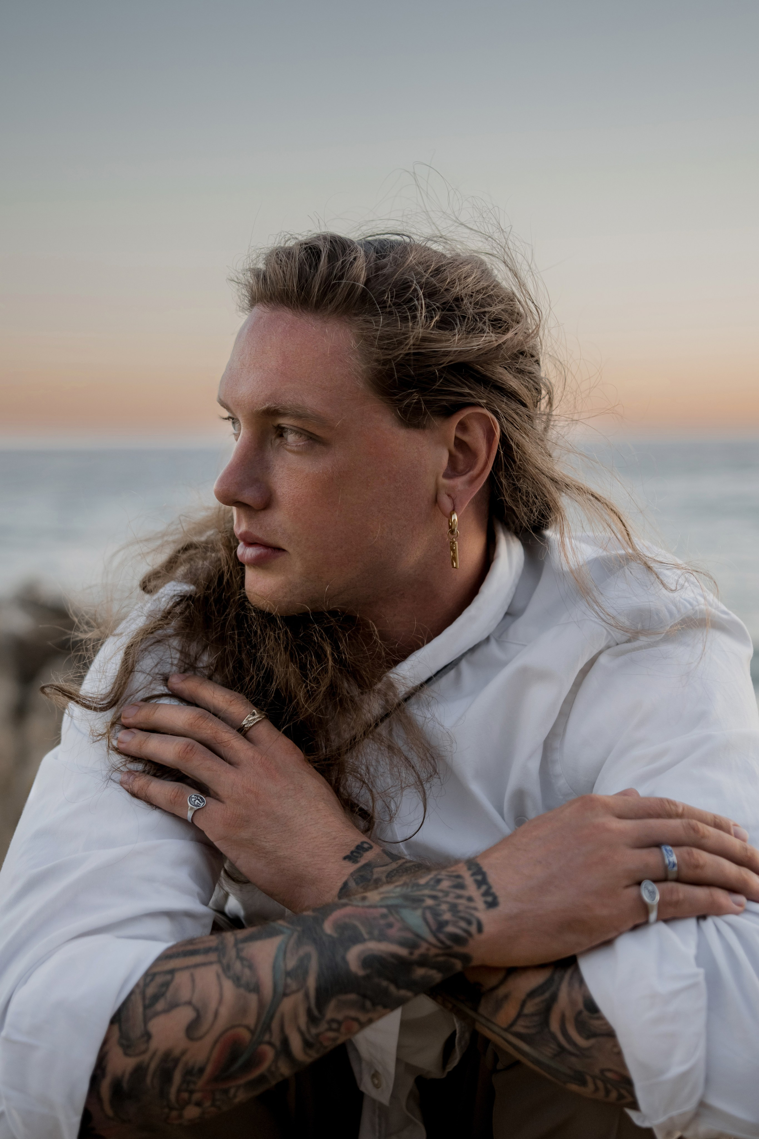 Male model wearing silver jewelry and casual attire, captured during golden hour by the ocean.