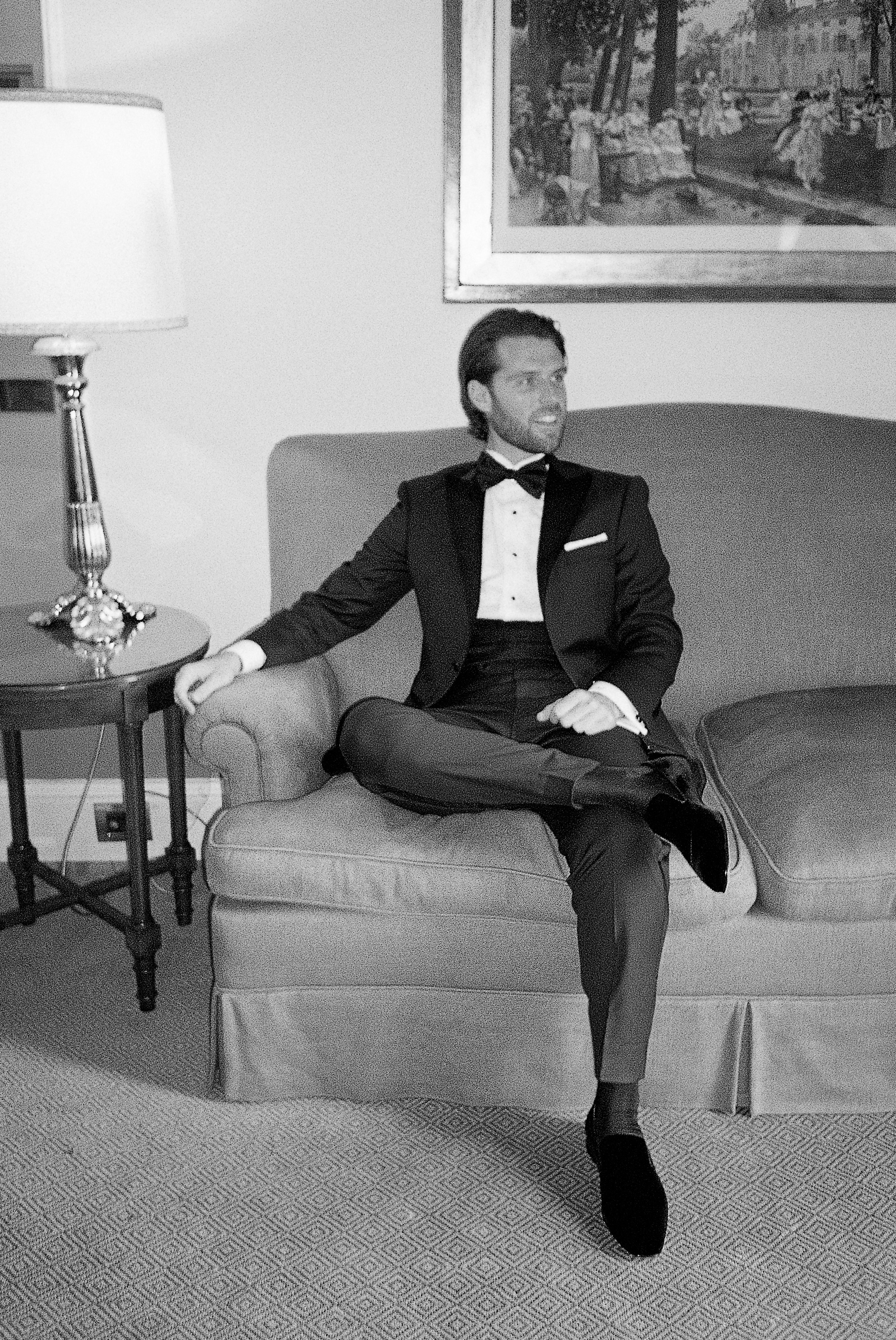 Groom in a classic tuxedo sits relaxed on a sofa, exuding elegance and calm sophistication in vintage villa interior. 