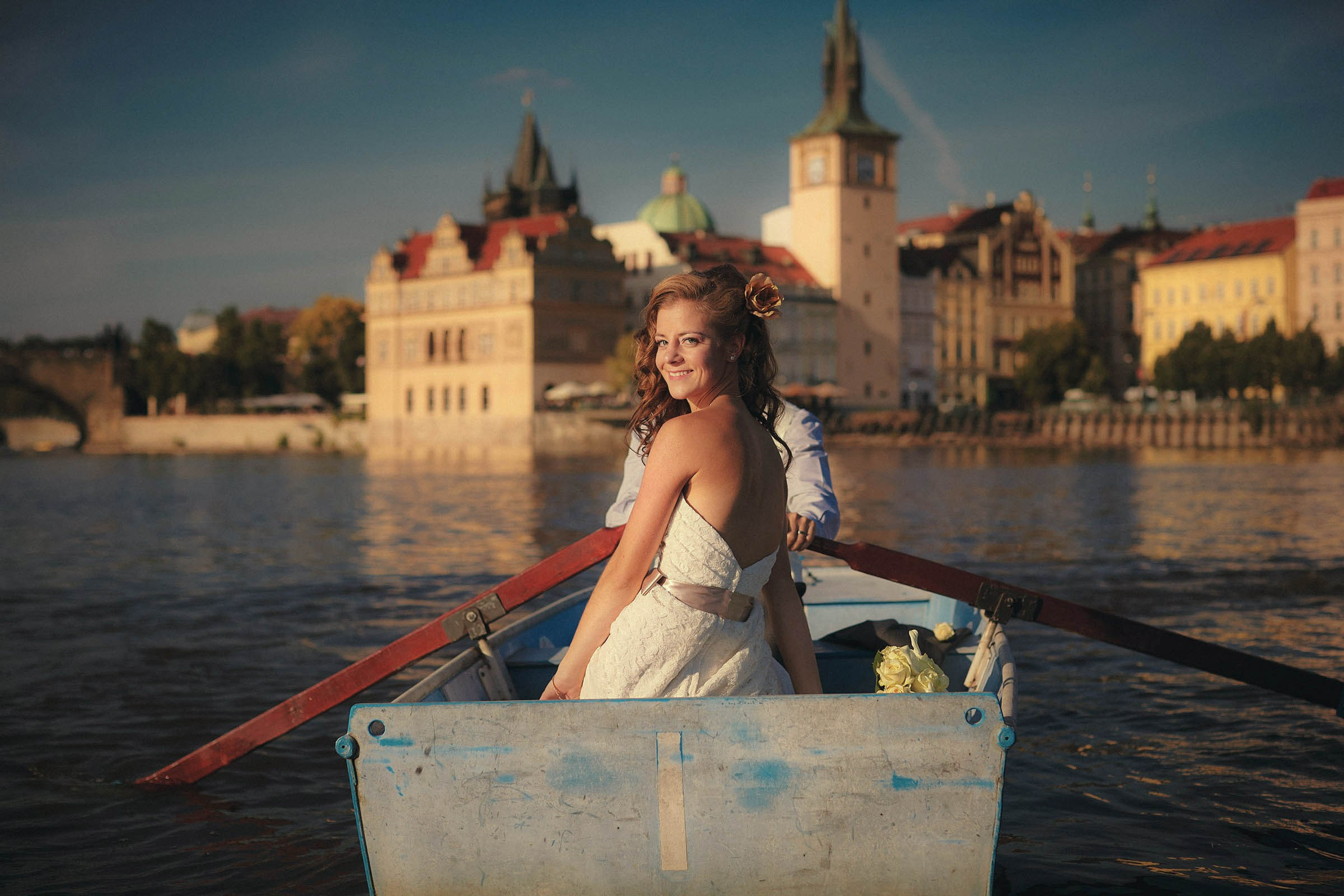 American bride looking at camera as groom rows boat on Vltava River, Prague.