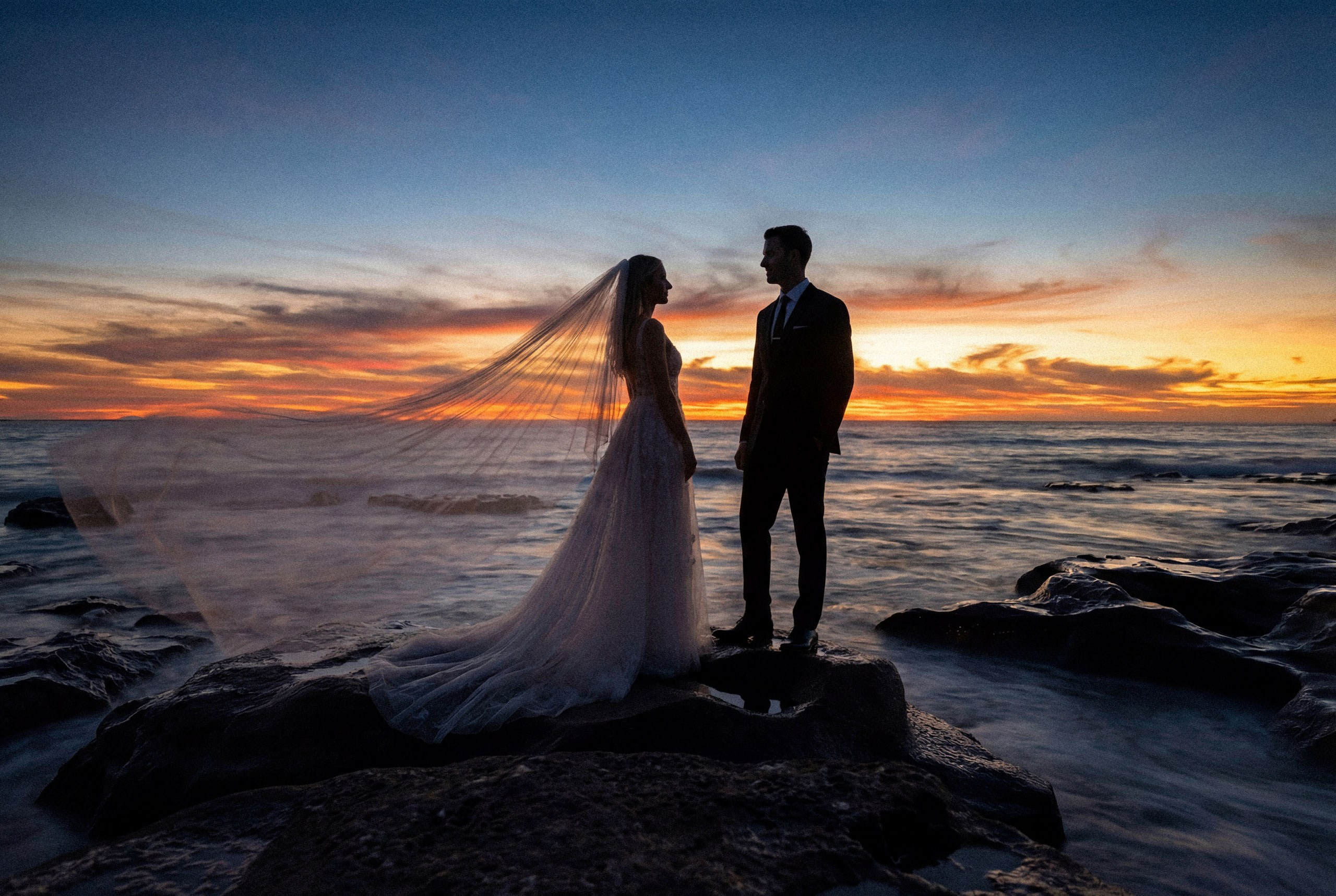 A romantic and dramatic photo of a bride and groom sharing a moment on a rock by the sea, their figures outlined by the fiery colors of the setting sun.