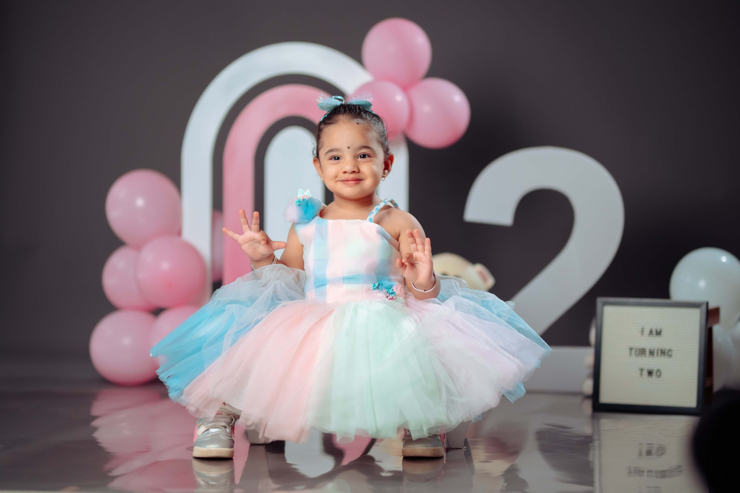 Baby girl in a white dress celebrating her birthday with a pastel balloon backdrop at Poudel Digital