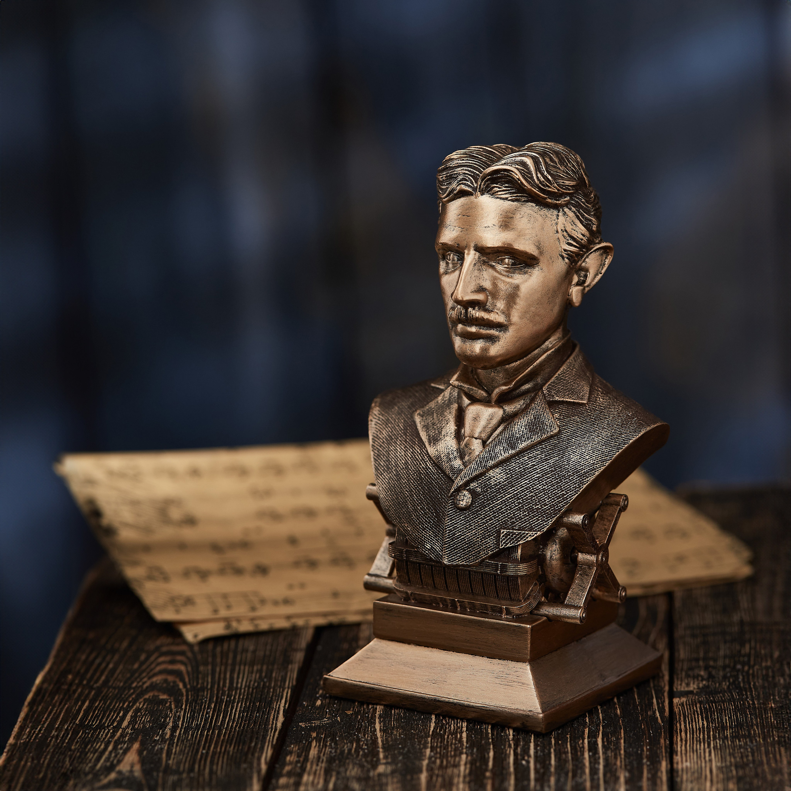 Nikola Teslanın bürünc büstü, qalın kostyumda, qaranlıq fonda taxta masa üzərində köhnə yazı kağızları yanında.