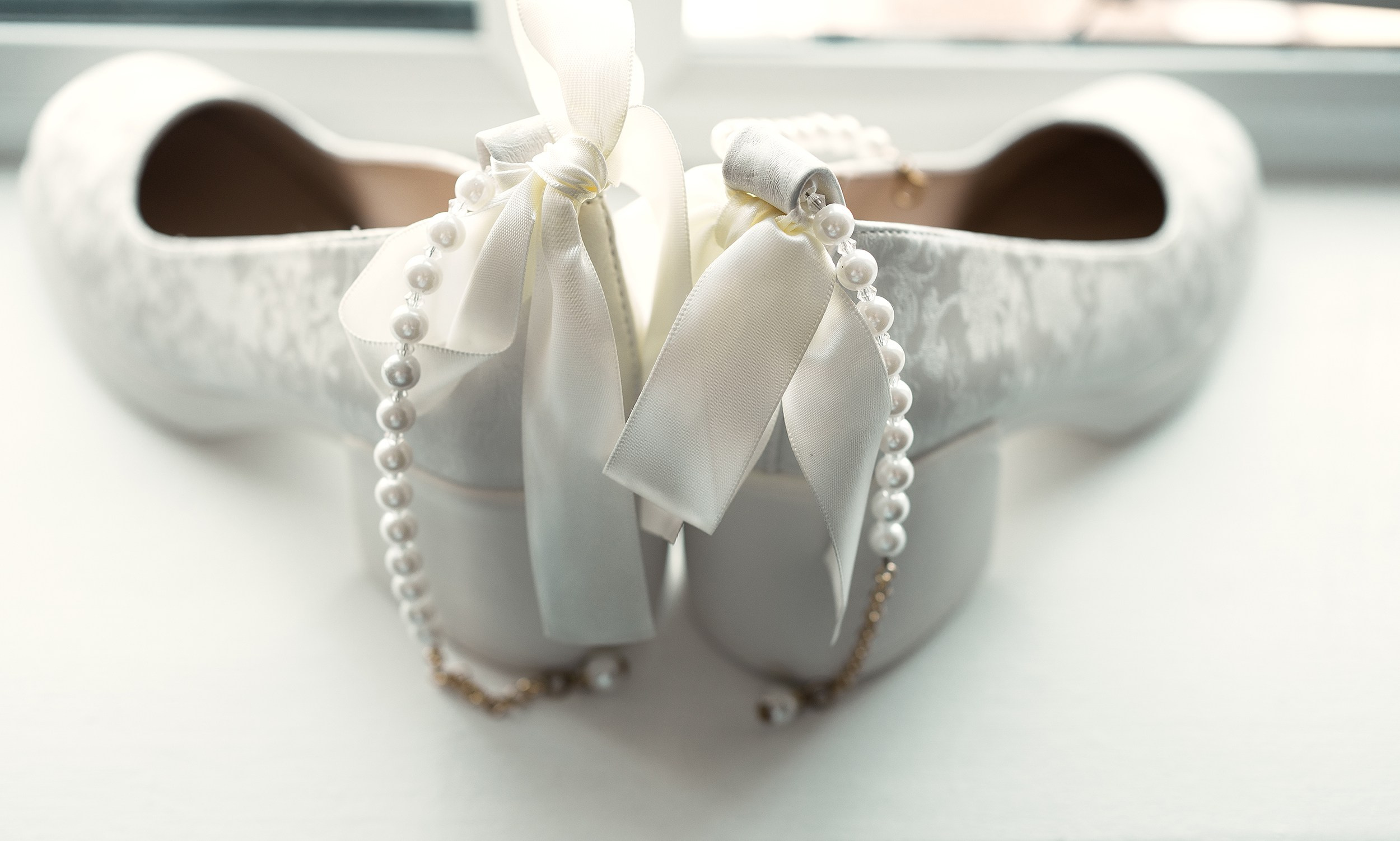 Close-up detail shot of elegant white bridal wedding shoes adorned with pearls and ribbons. Professional wedding photography details by vdphotoart.