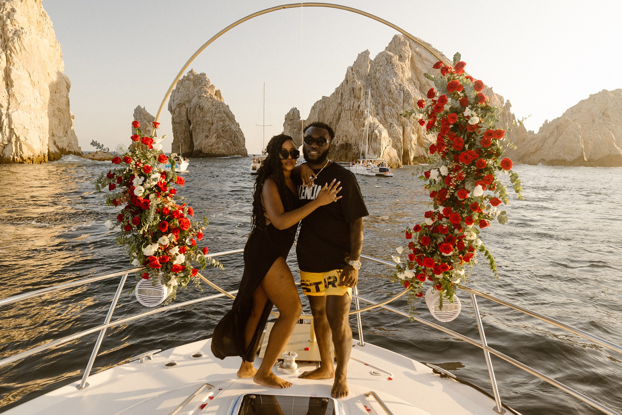 Yacht engagement in Cabo San Lucas – romantic Afro couple with red floral arrangement and El Arco in background, luxury proposal photographer Los Cabos