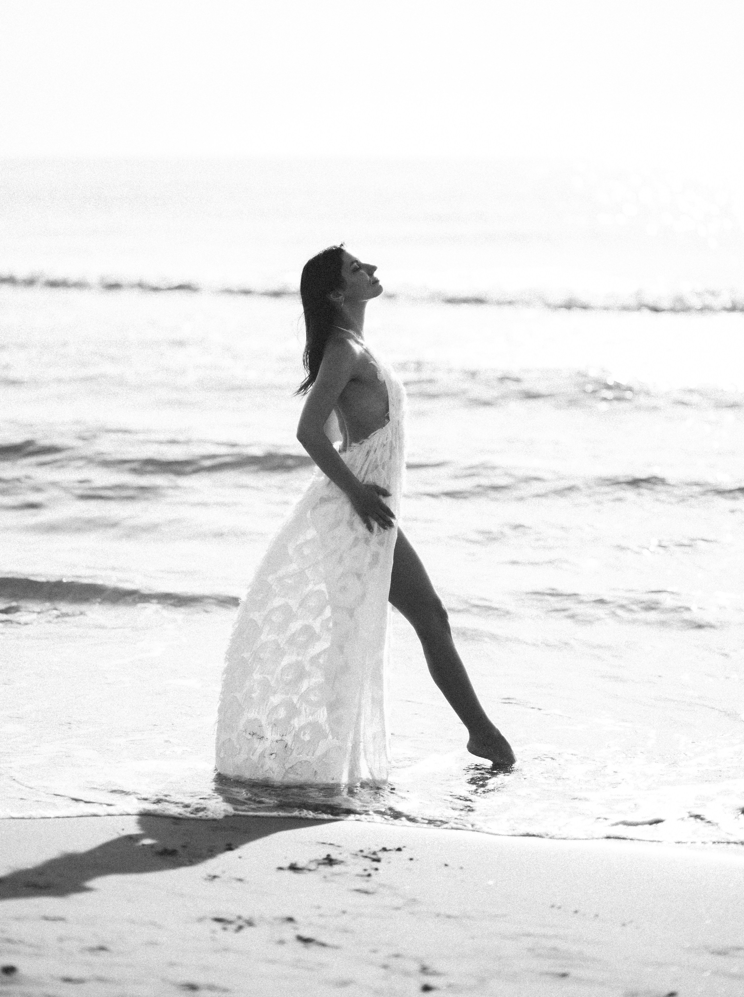 Грациозная женщина в воздушном полупрозрачном платье позирует босиком на берегу во время заката в Кальпе, Испания — художественная черно-белая пляжная портретная фотосъемка, передающая элегантность, движение и атмосферу Средиземного моря, идеально подходящая для портретных фотосессий в Кальпе и Испании.