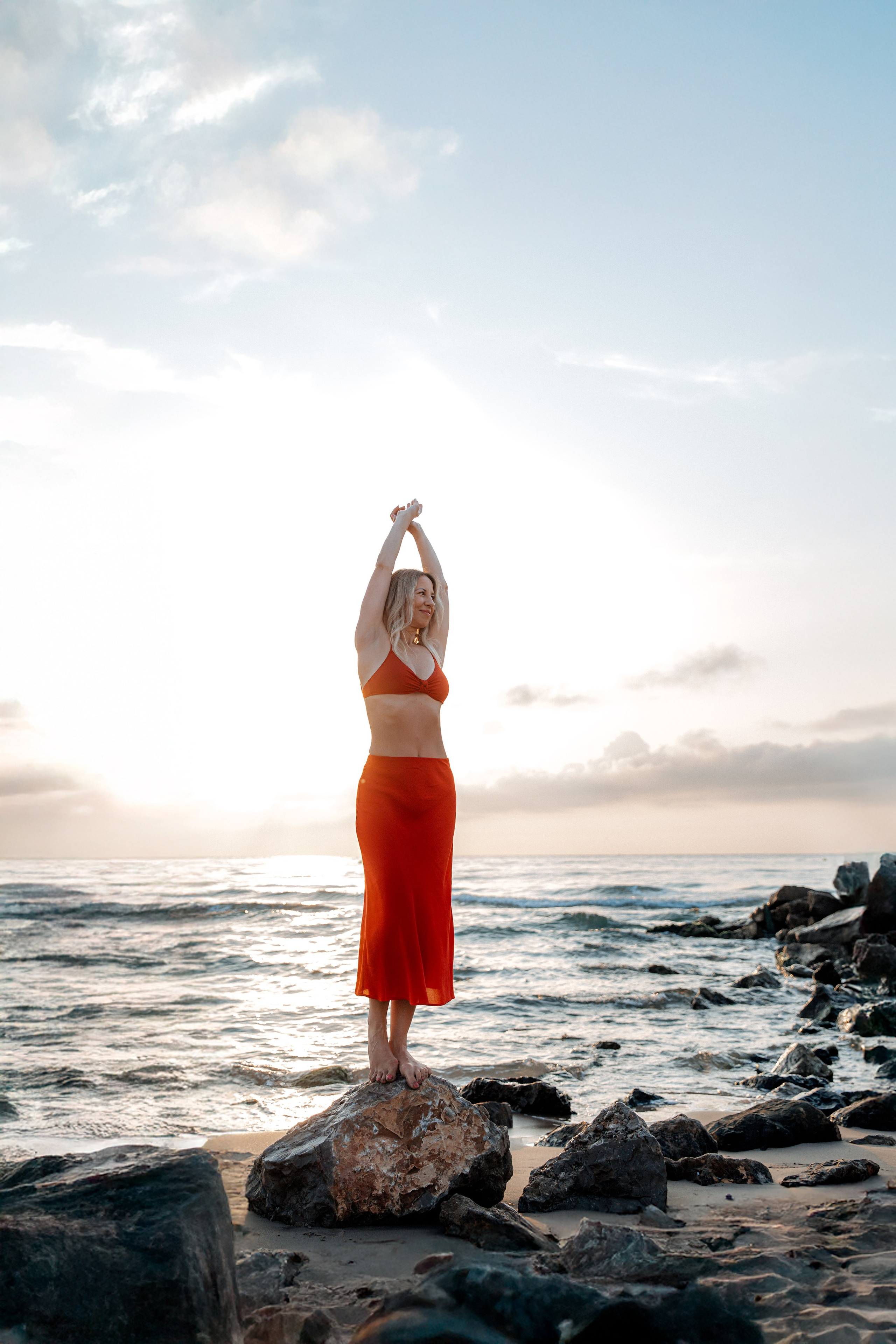 Улыбающаяся женщина в красном наряде стоит босиком на прибрежных скалах на рассвете в Кальпе, Испания — яркая пляжная портретная фотография, передающая радость, естественный свет и красоту Средиземноморского побережья, идеально подходит для портретных фотосессий в Кальпе и Испании.