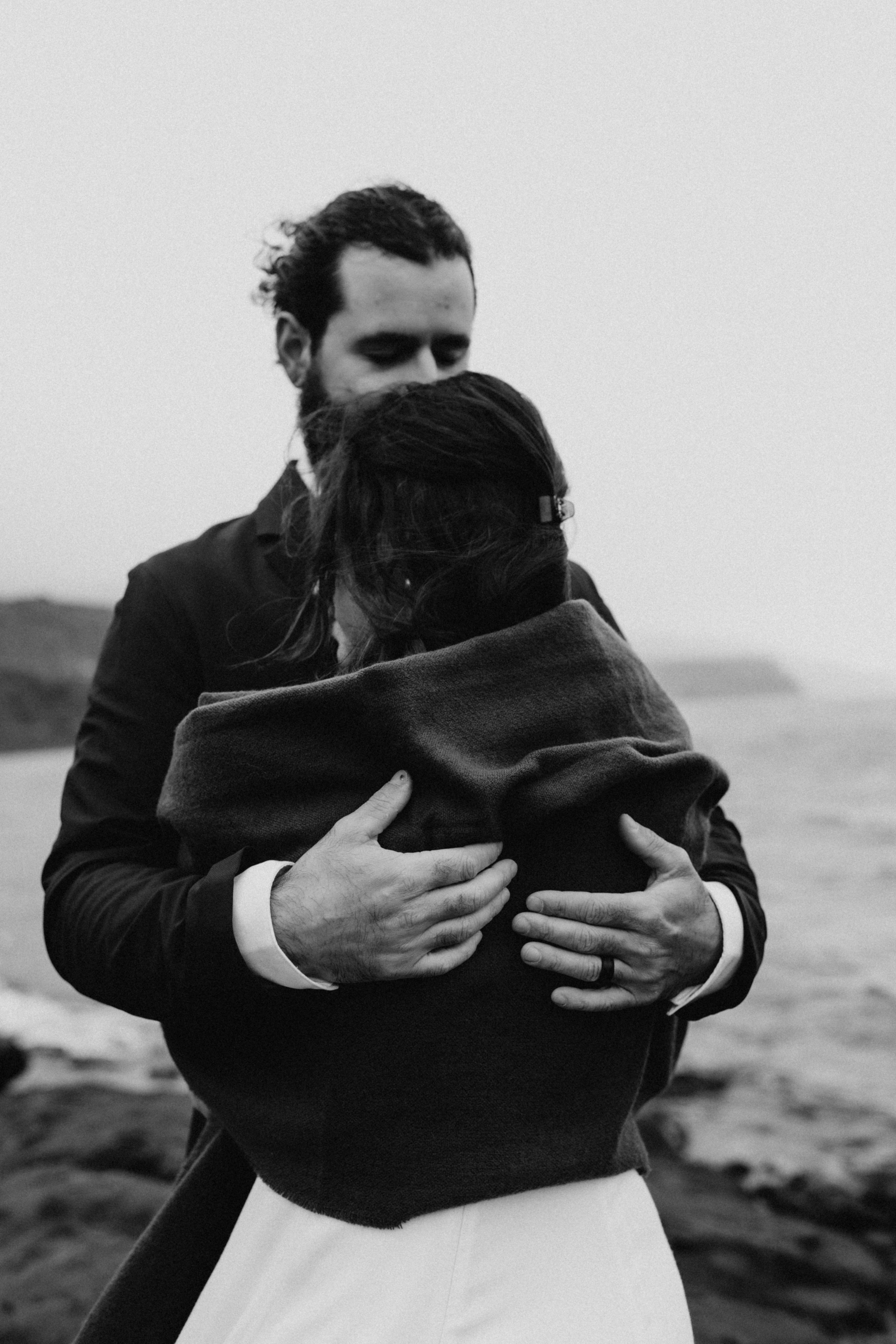 Groom wrapping his arms around the bride as they stand on the rocky edge of Kleifarvatn, braving the cold Icelandic wind.