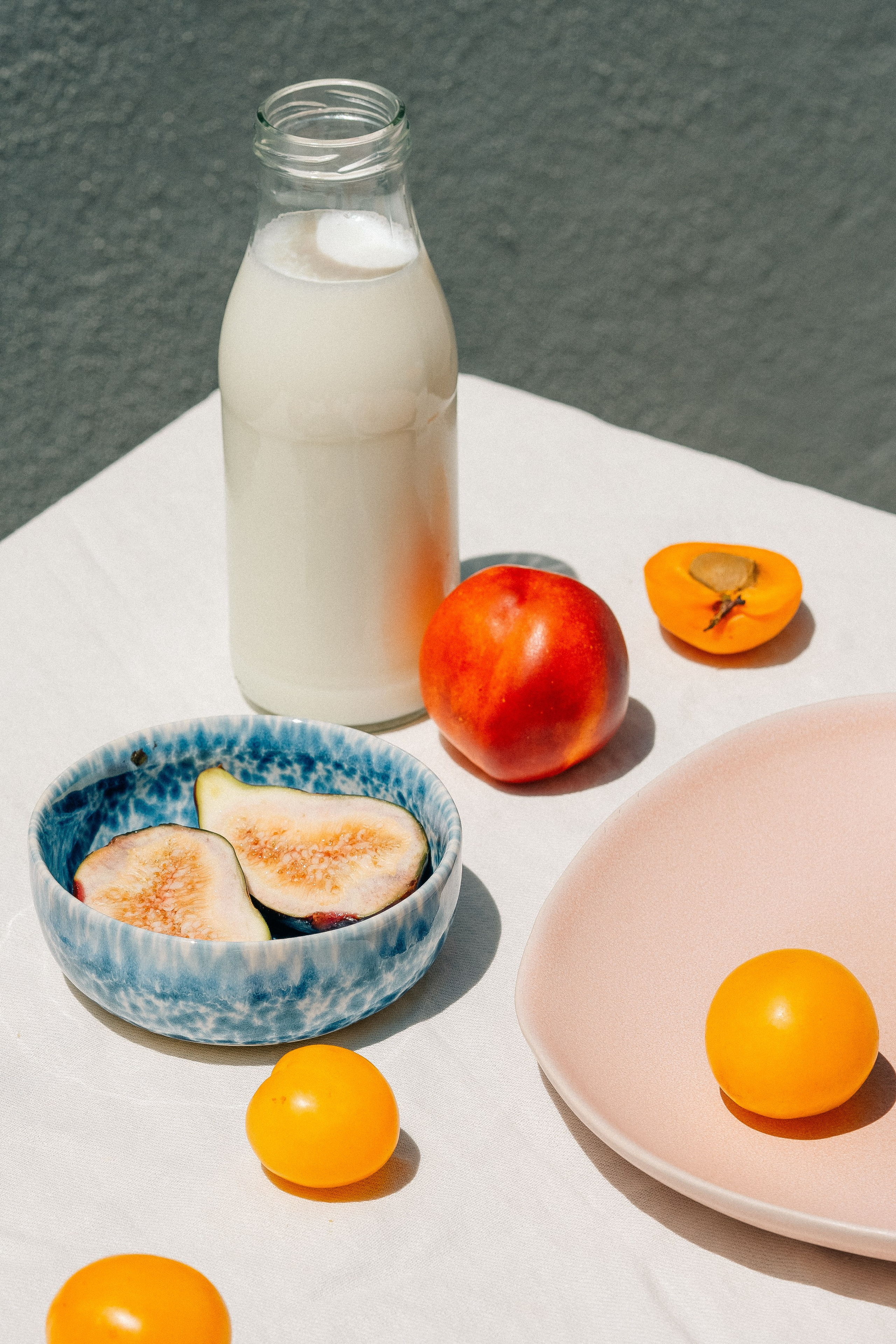Fruit on plates with a bottle of milk. Peaches, apricots, and figs lying on a table and plates. The plates are blue and pink. A bottle of milk stands near fruit. All objects stand on a table covered with a white. 
