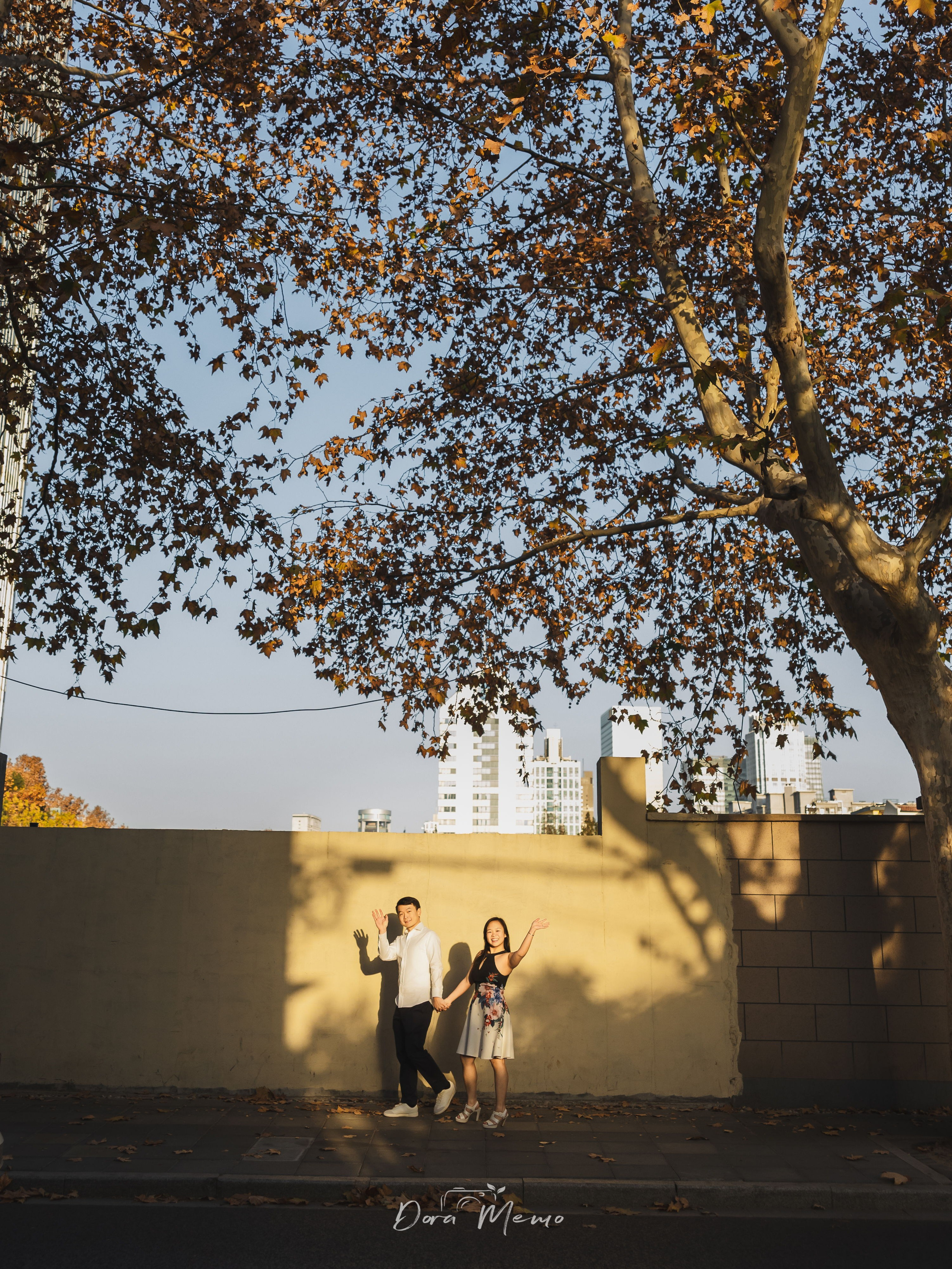 Couple waving beneath a large tree with long afternoon shadows, quiet closing