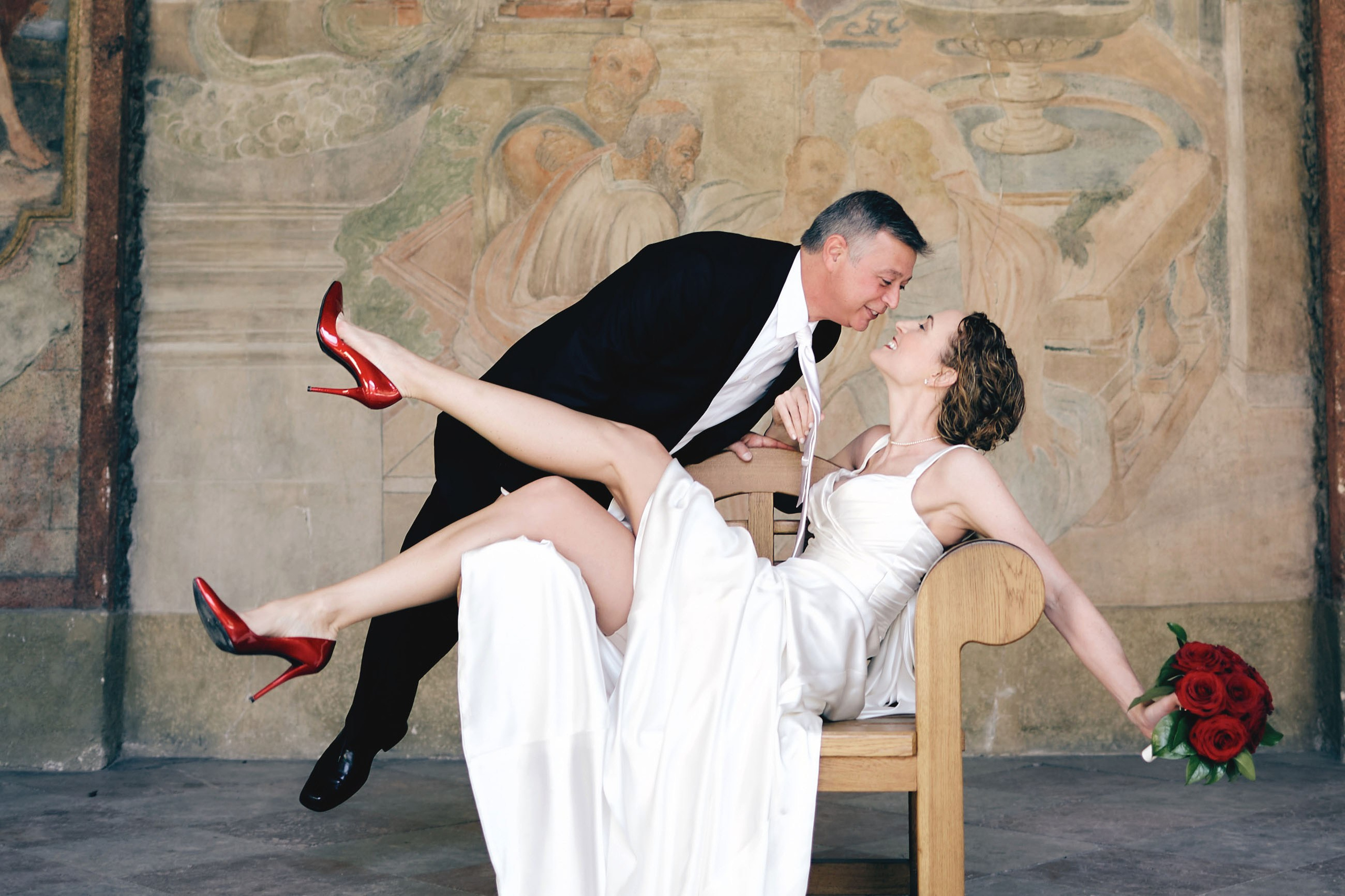 Bridal charm at Vrtba Garden: bride in red pumps pulling groom by tie, seated in stylish chair, Prague.