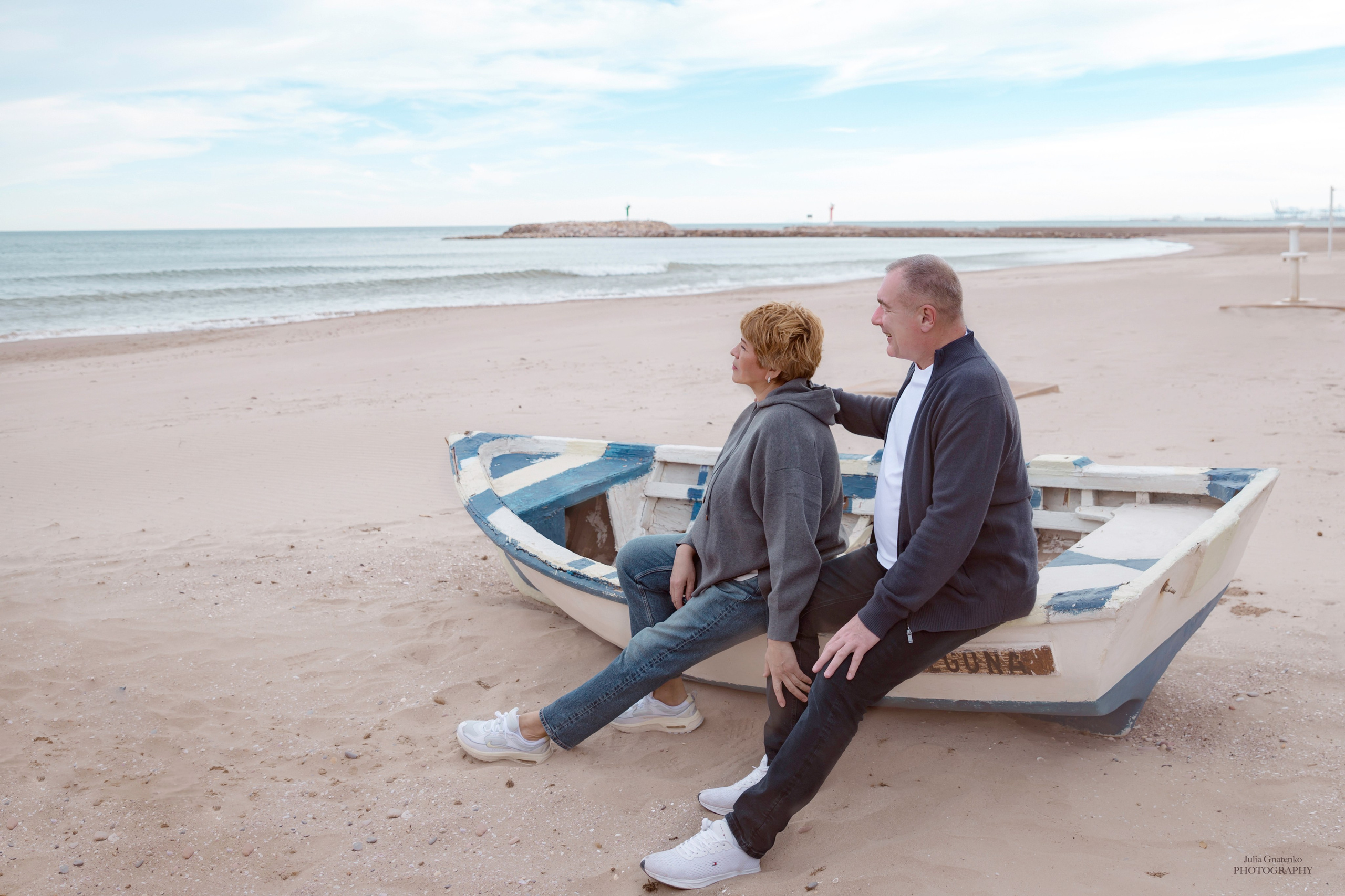A romantic moment captured as two men sit closely together on a rustic wooden boat along a quiet beach, gazing toward the sea under soft skies in Valencia, Spain.
