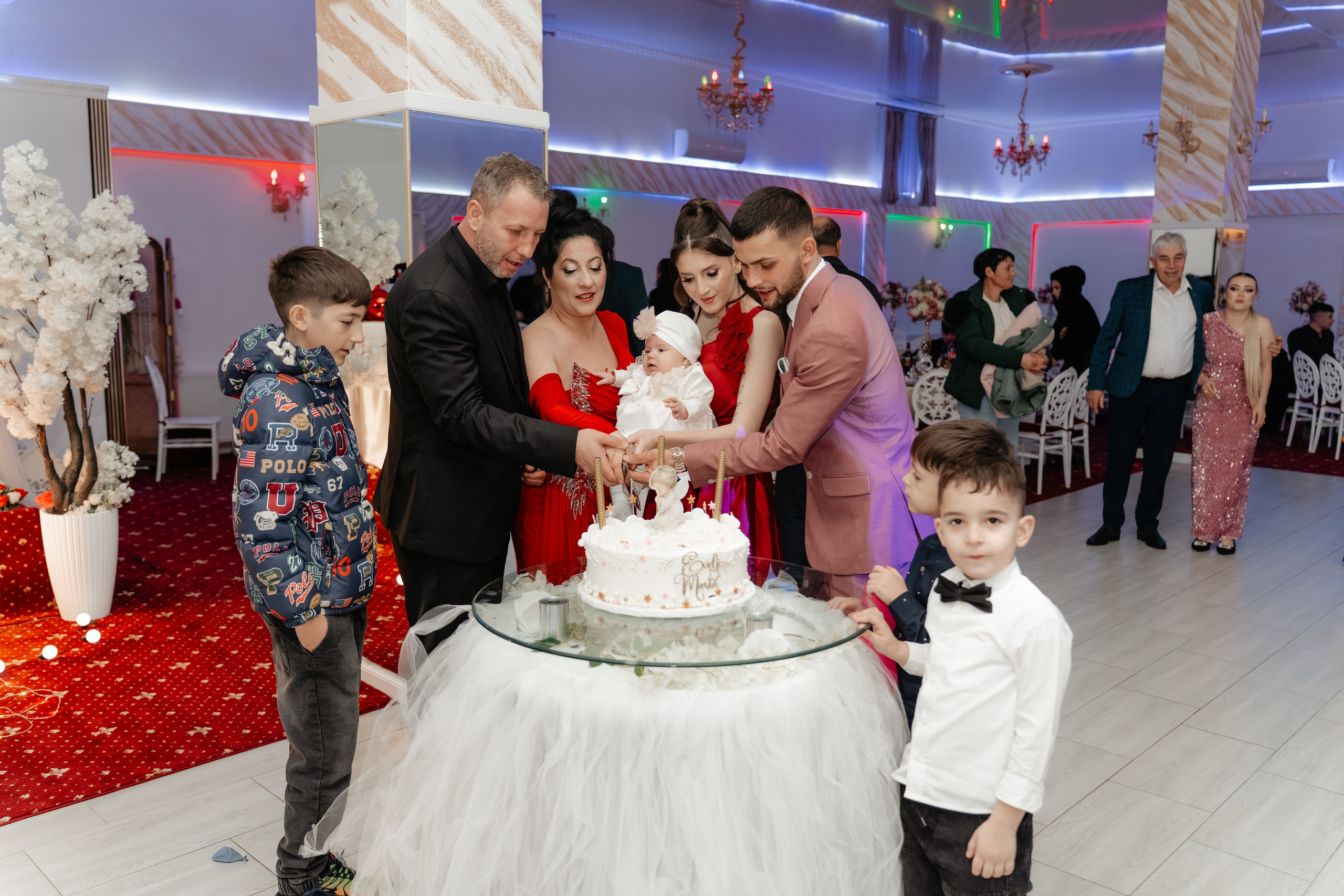 Cadru lateral intim al tăierii tortului: familia și nașii, concentrați și bucuroși, taie primul felie din tortul de botez al Evelinei Maria.