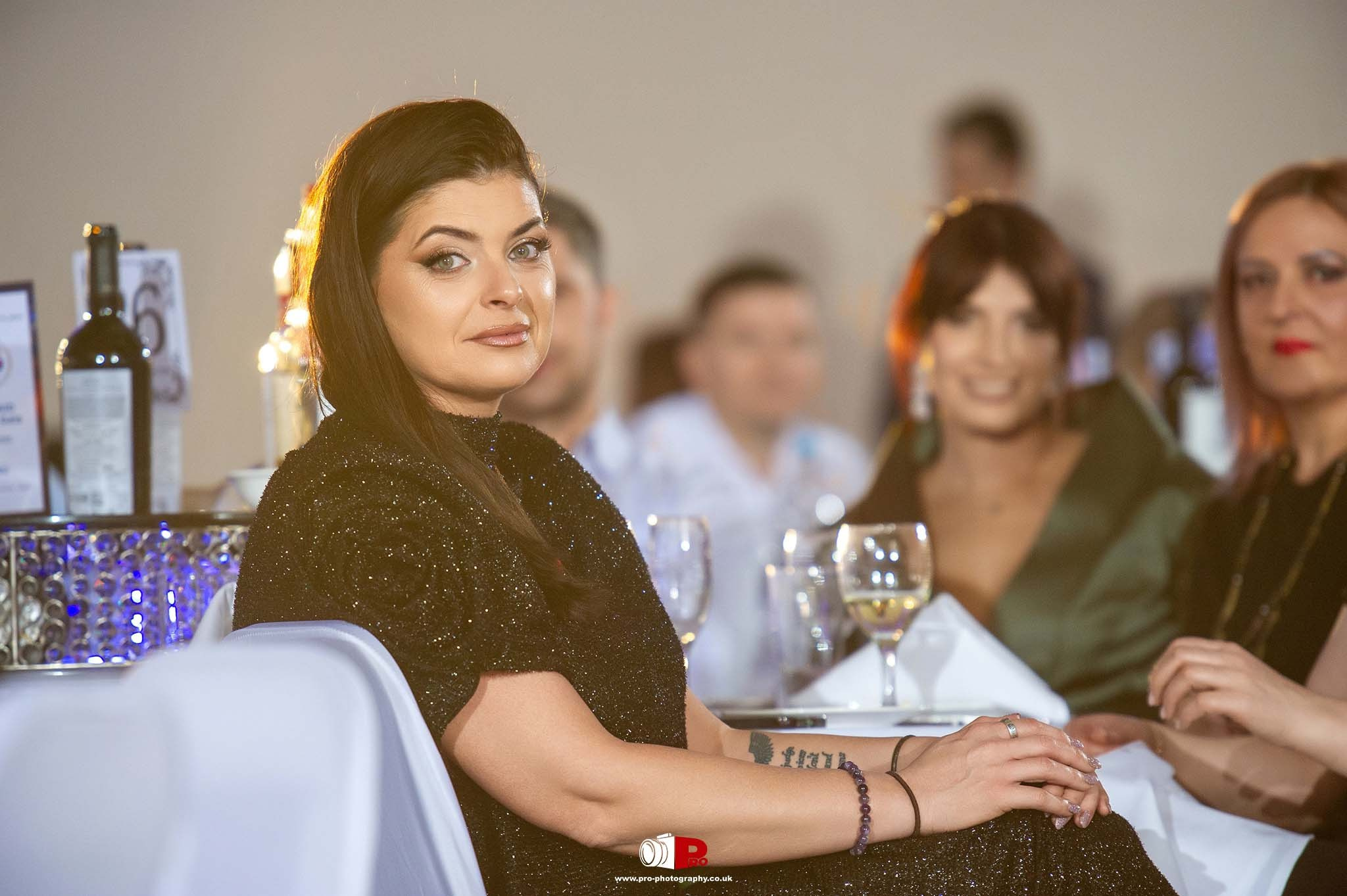 Andreea Hasie, in a black glittering dress, smiles during a formal dinner event. Wine glasses are on the table.