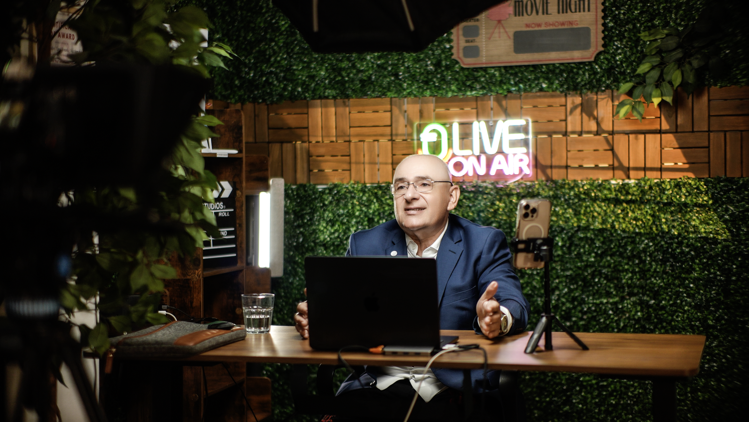 Podcast and livestream set at ProStudio1, Hollywood Florida — host at a desk with microphone and “LIVE ON AIR” sign in a greenery backdrop.